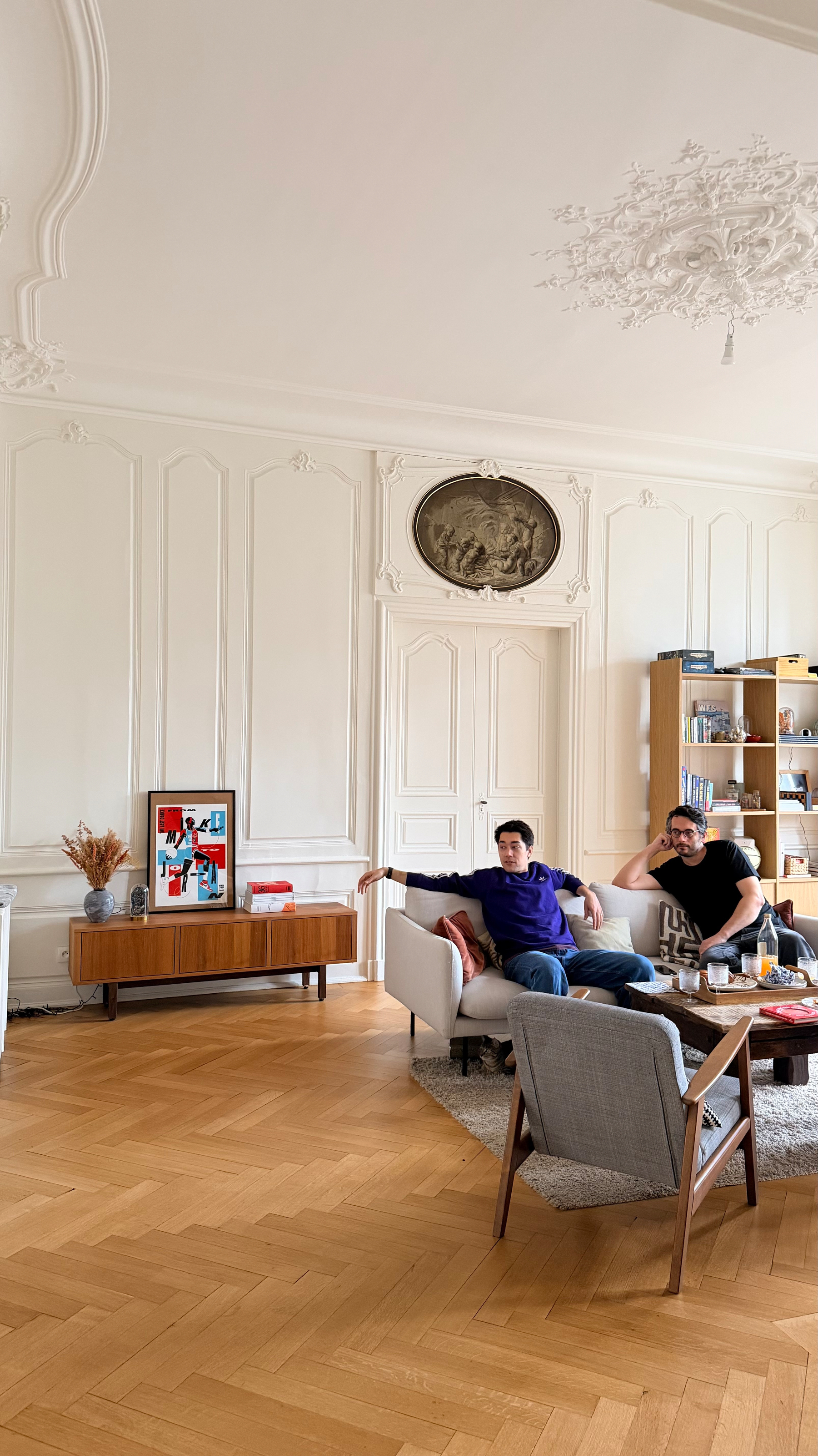 Two people are sitting on a couch in a spacious living room with elegant white walls, wooden floors, and bookshelves.