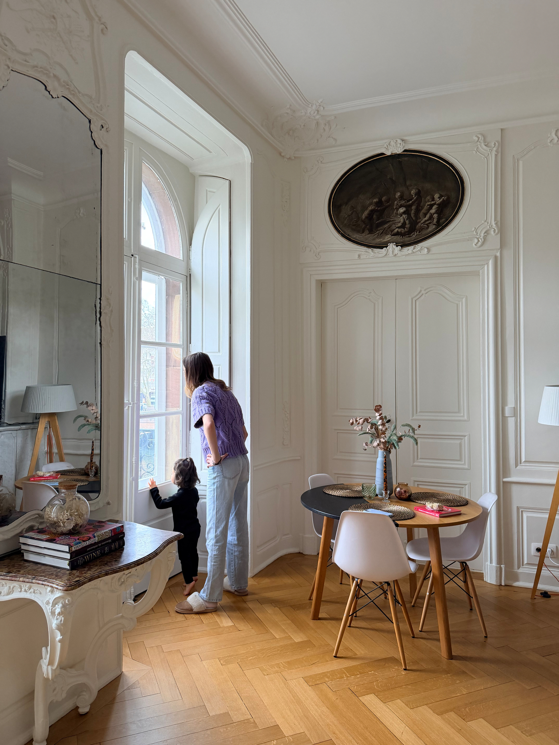 A woman and a child are looking out a tall window in a cozy, elegantly decorated room with a small dining table and chairs.
