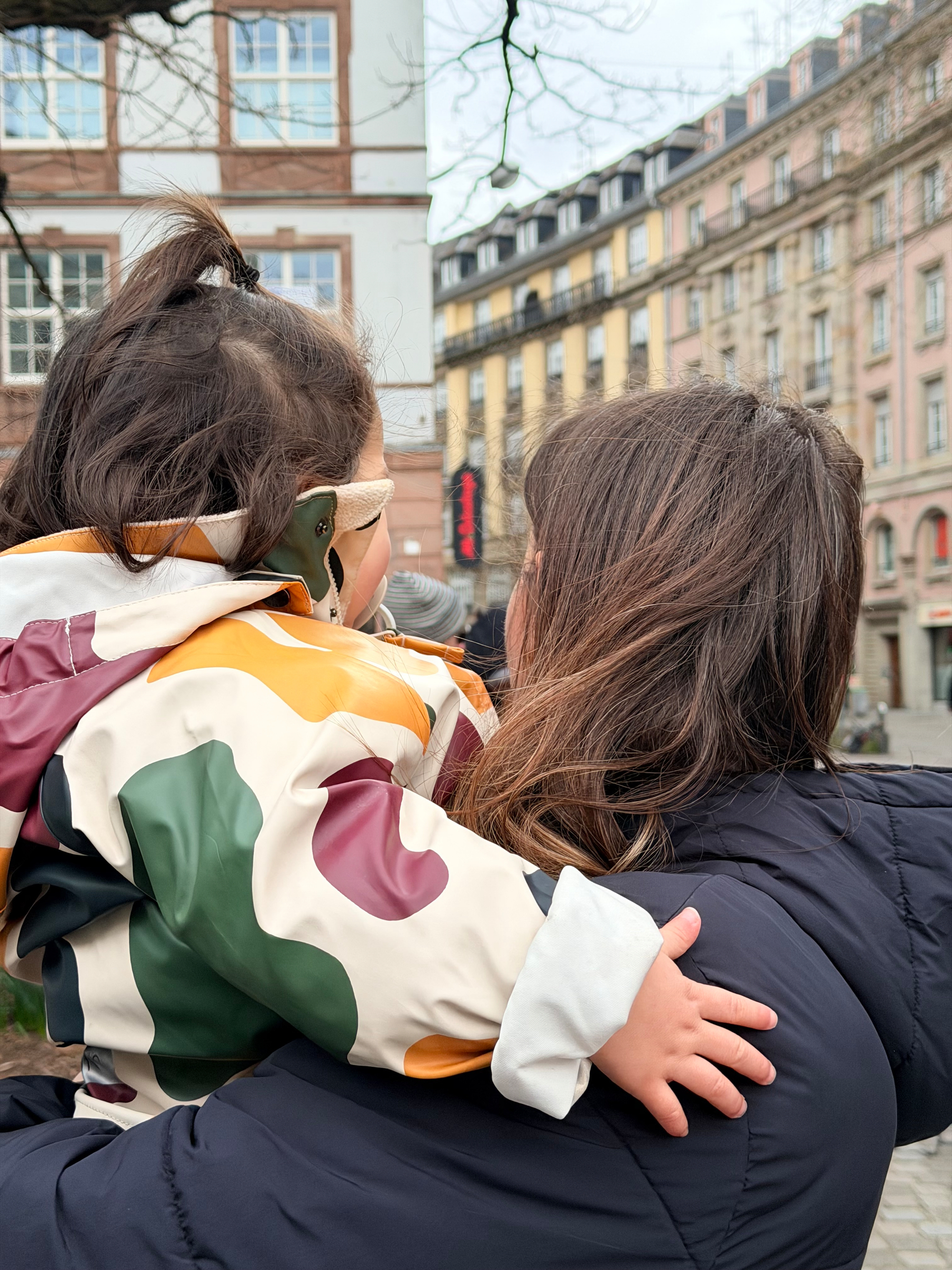 A woman is holding a child while standing in an urban area with buildings in the background.