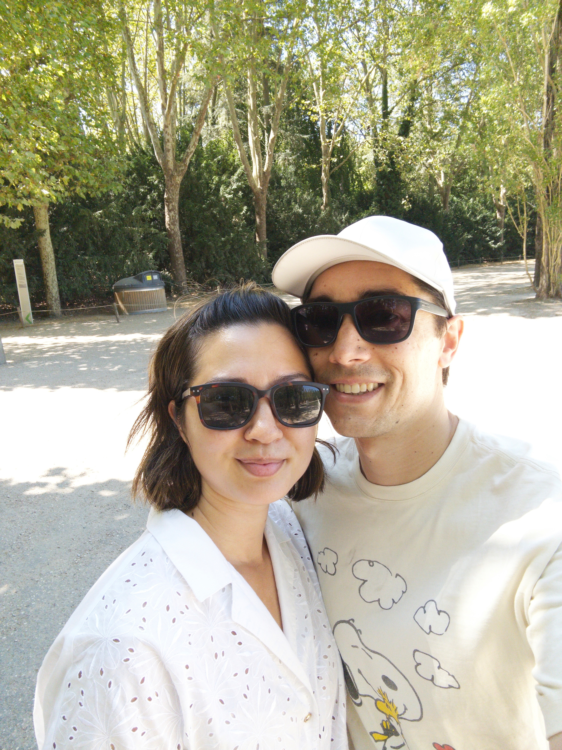 Auto-generated description: A smiling couple wearing sunglasses poses for a selfie outdoors on a sunny day with trees in the background.