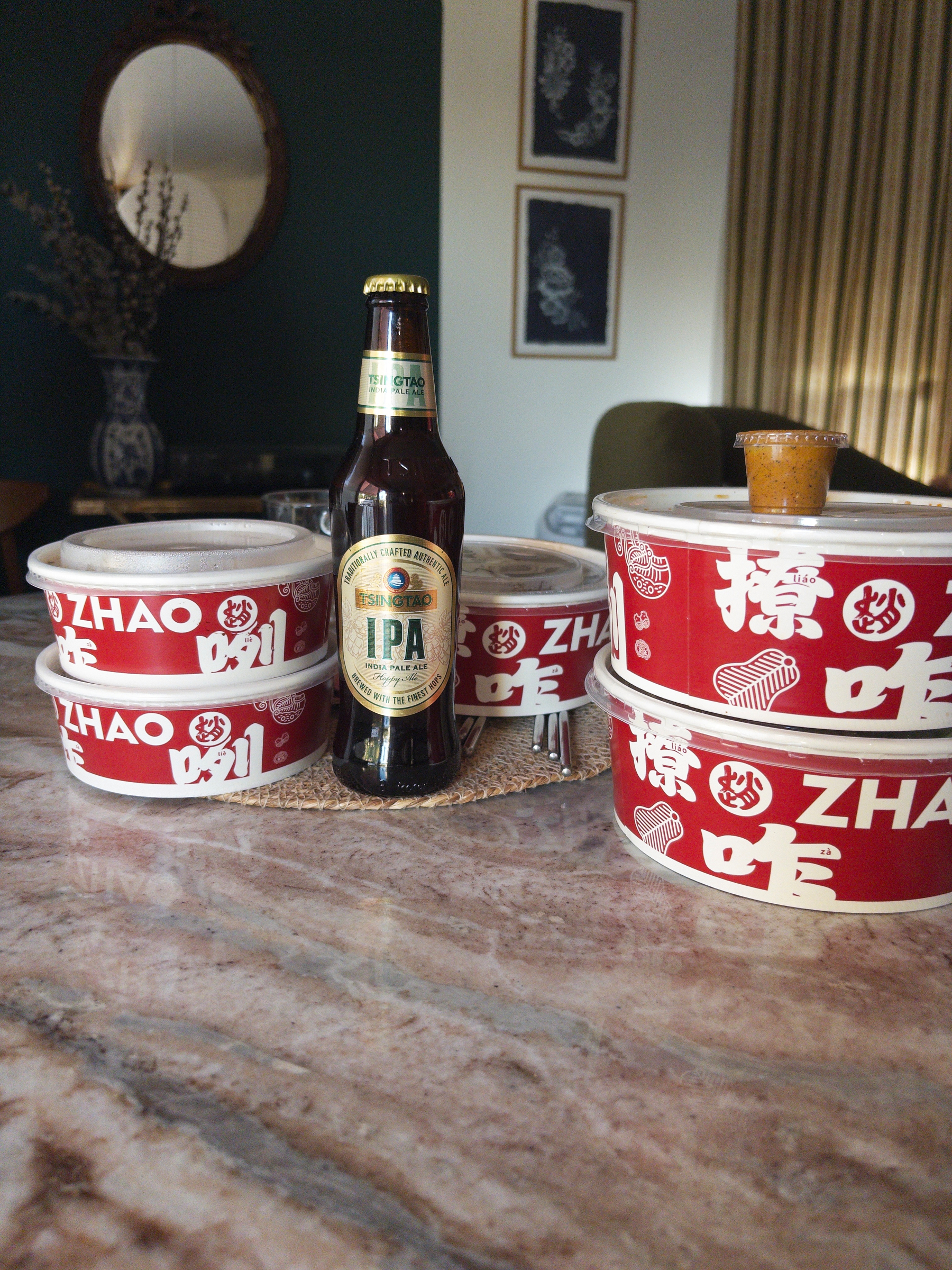 Auto-generated description: A bottle of Tsingtao IPA beer is placed among several stacked takeout containers with red and white labeling on a marble table.