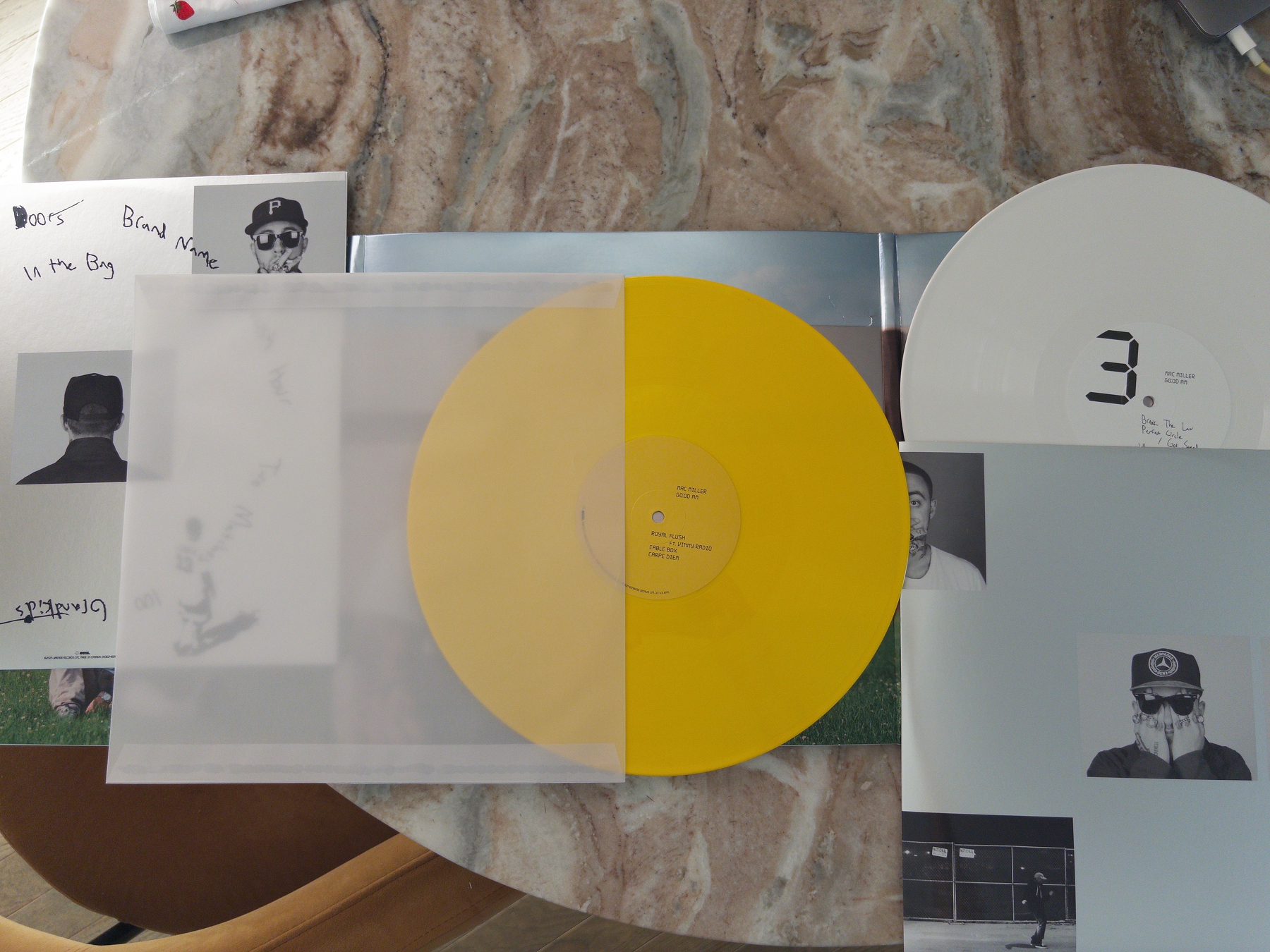 Two vinyl records, one yellow and one white with a large number 3 on it, are displayed on a marble table along with album covers and inserts featuring artist portraits and track listings.