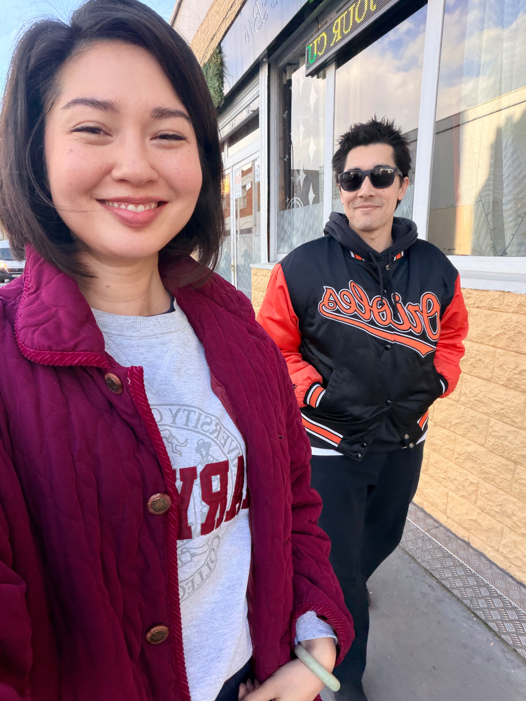 Auto-generated description: Two people are smiling outdoors, with one wearing a burgundy jacket and the other a black and red jacket.