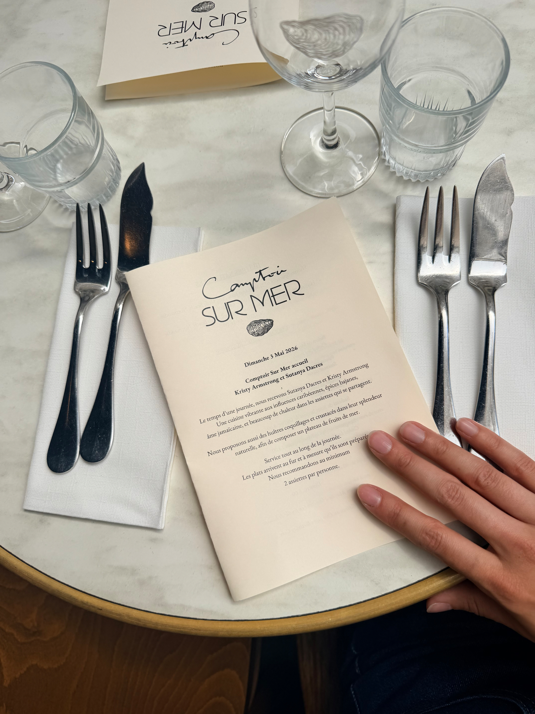 A hand is holding a menu from Comptoir Sur Mer on a table set with cutlery, glasses, and napkins.