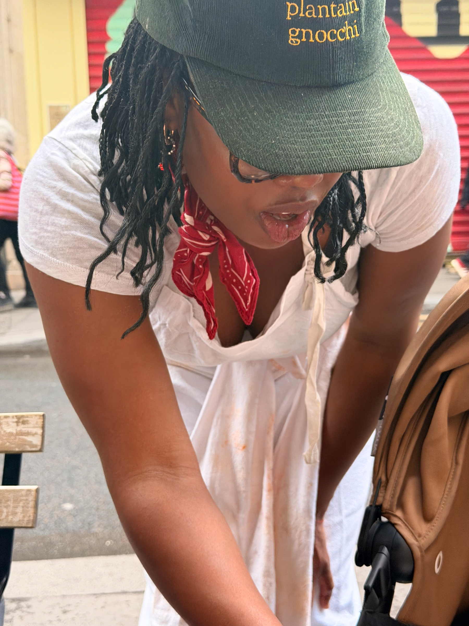 A person wearing a green cap, red bandana, and white dress is leaning over a stroller on a street.
