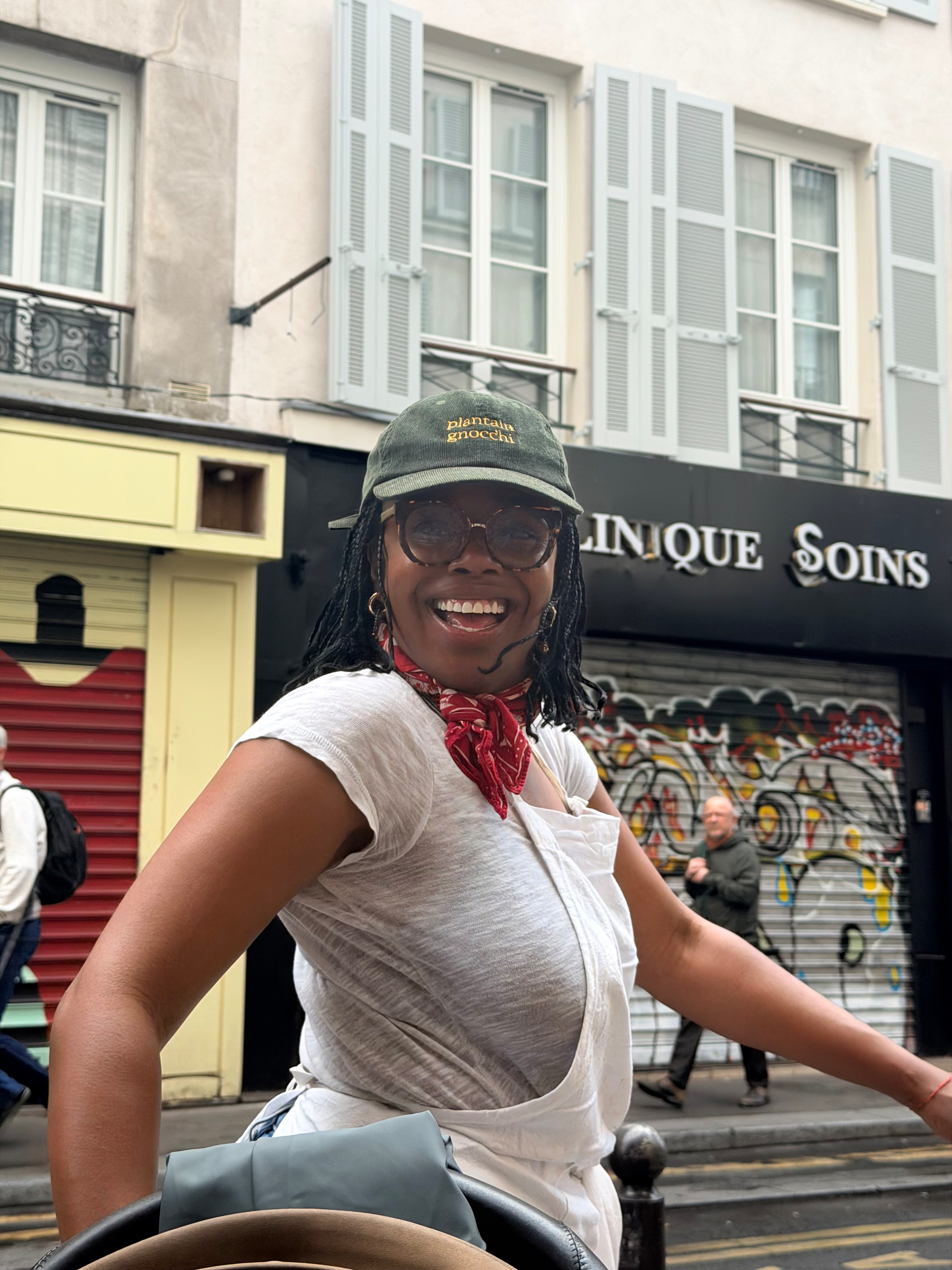 A person wearing glasses, a cap, and a red bandana is smiling while standing on a city street.