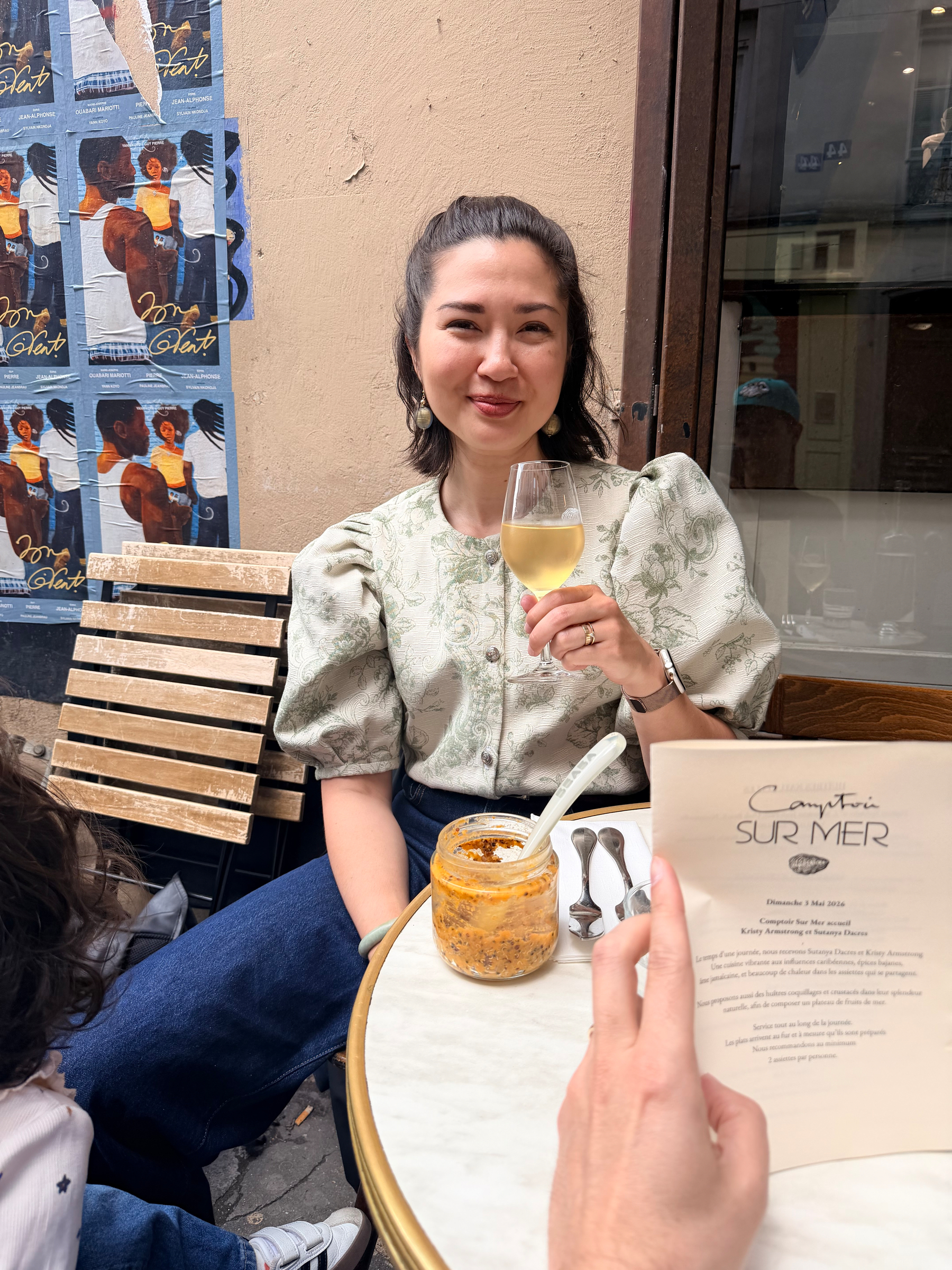 A woman is sitting at an outdoor cafe table, holding a drink and smiling, with another person holding a menu in the foreground.
