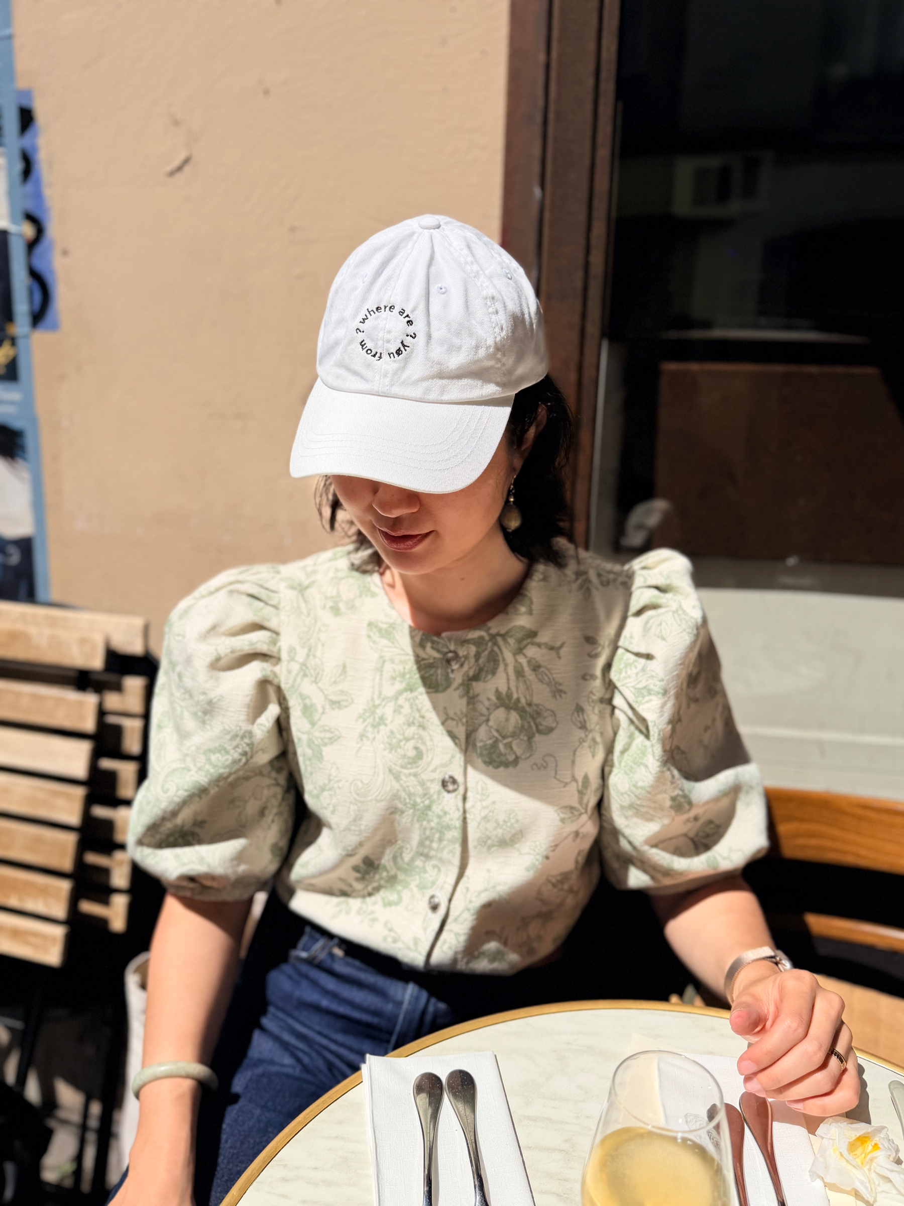 A person wearing a white cap and floral blouse is sitting at a sunlit outdoor café table set with utensils and a drink.