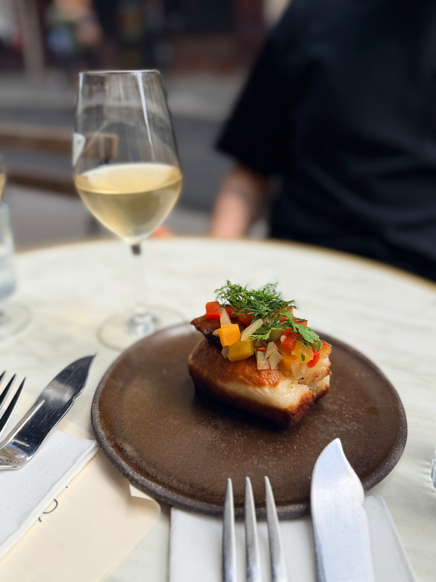 A plated dish of food topped with vegetables is on a table next to a glass of white wine and cutlery.