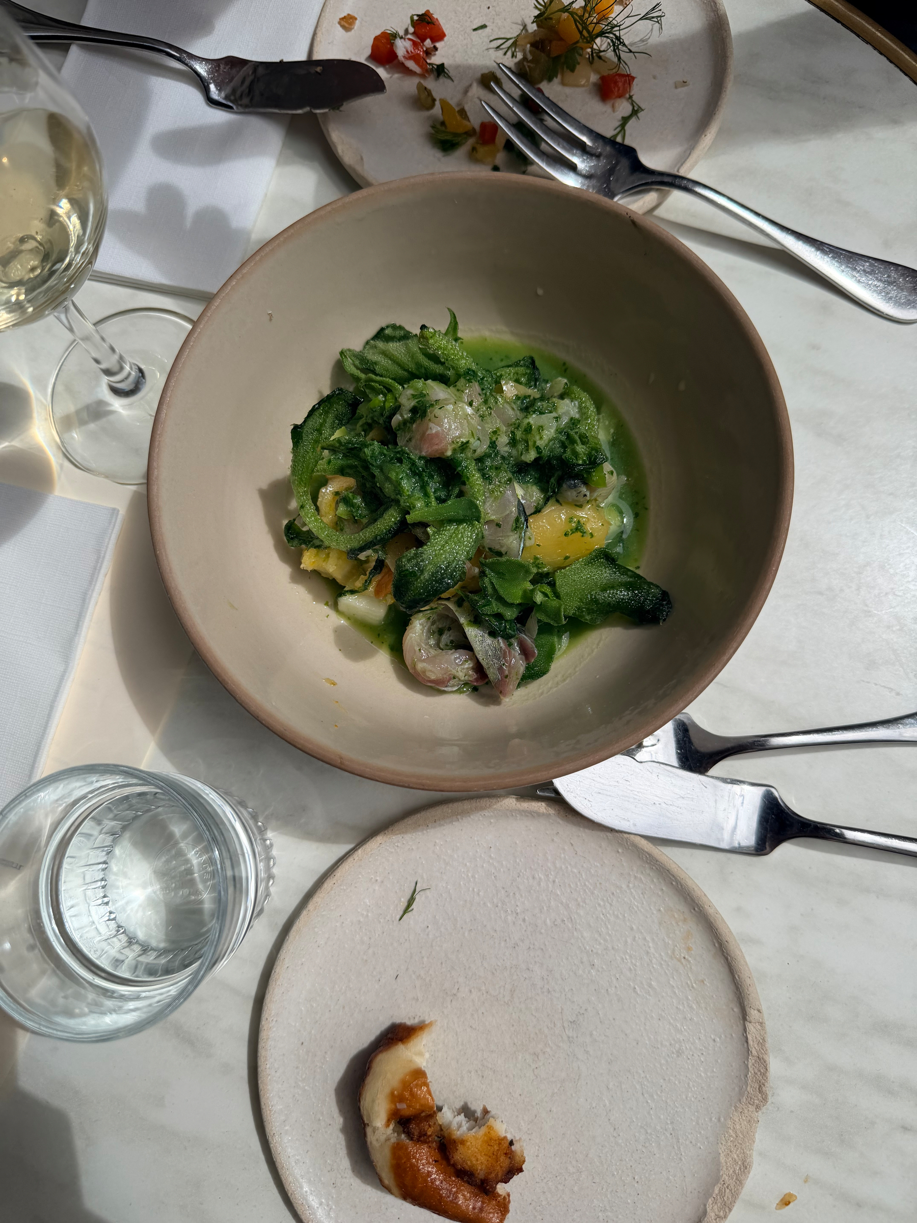 A table setting features a bowl of pasta with green sauce, garnished with herbs, alongside a piece of bread, a glass of white wine, and a glass of water.