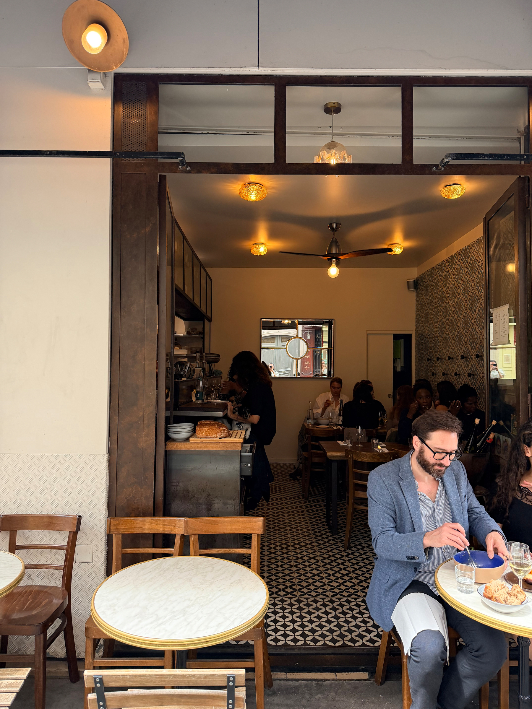 A cozy café scene with people enjoying food and drinks, featuring outdoor and indoor seating and warm lighting.