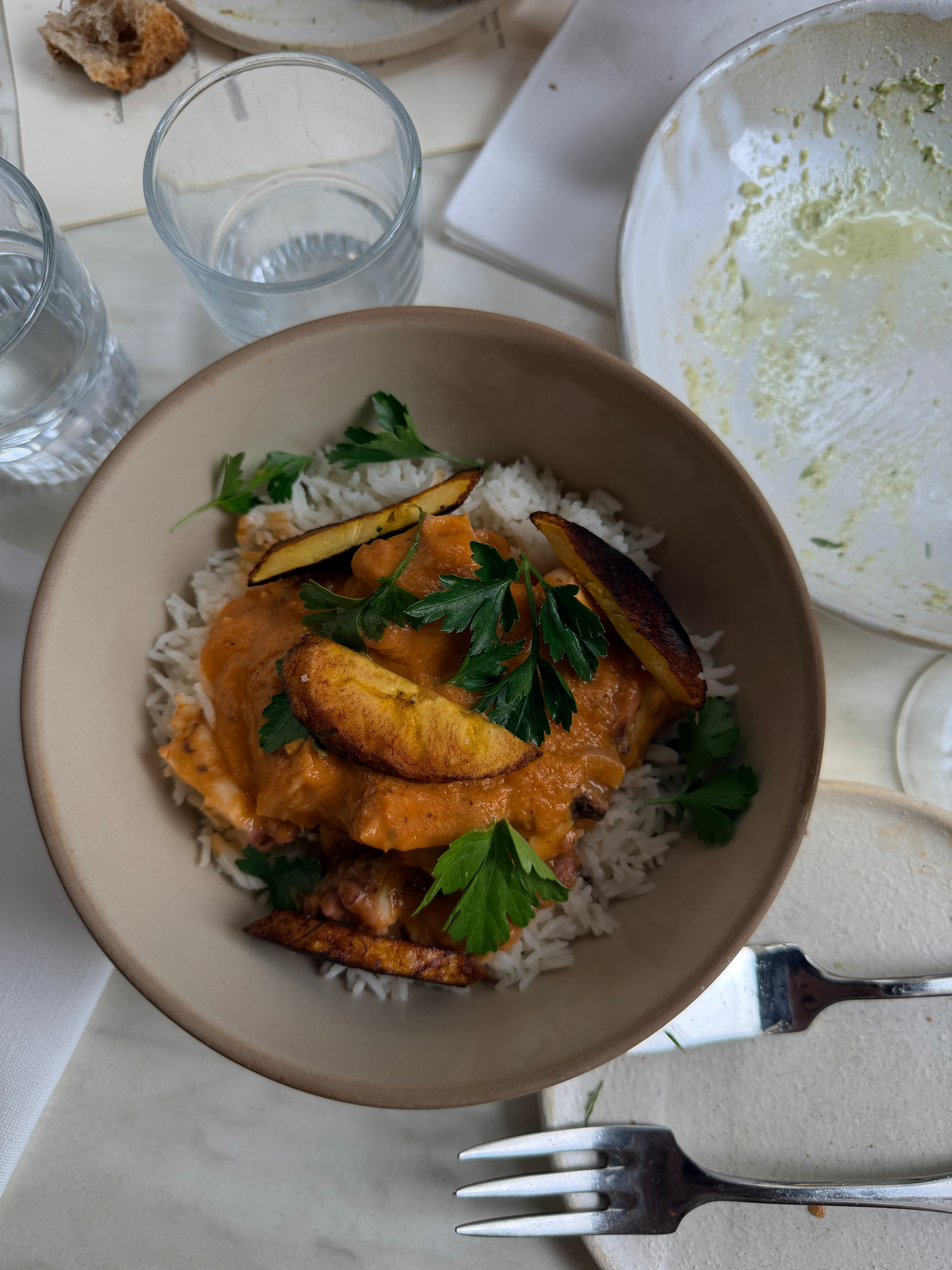 A dish featuring rice topped with sauce, plantains, and garnished with parsley is served in a bowl next to a partially eaten meal, a fork, a knife, and glasses of water.