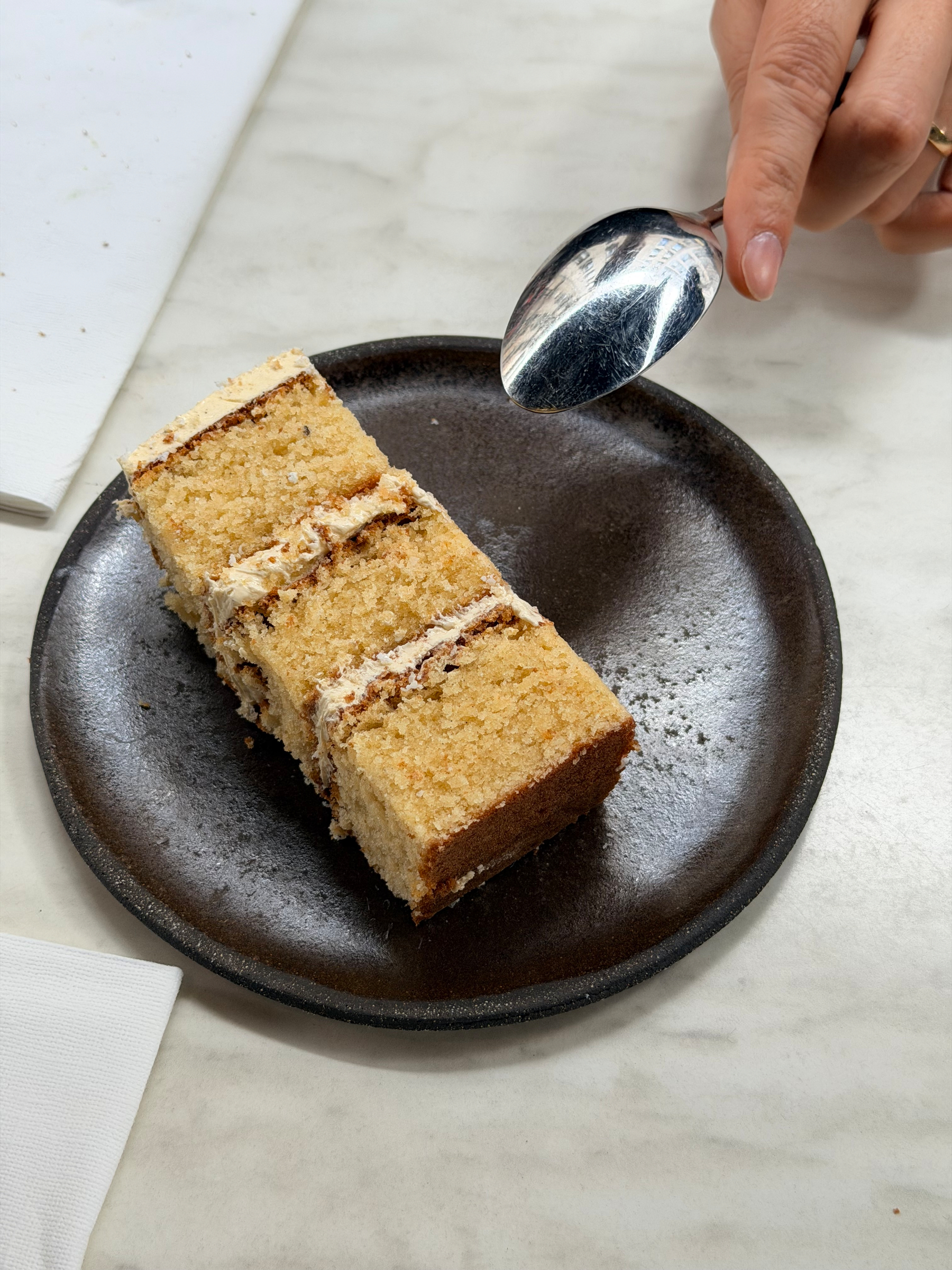 A slice of layered cake is on a dark plate, with a hand holding a spoon nearby.