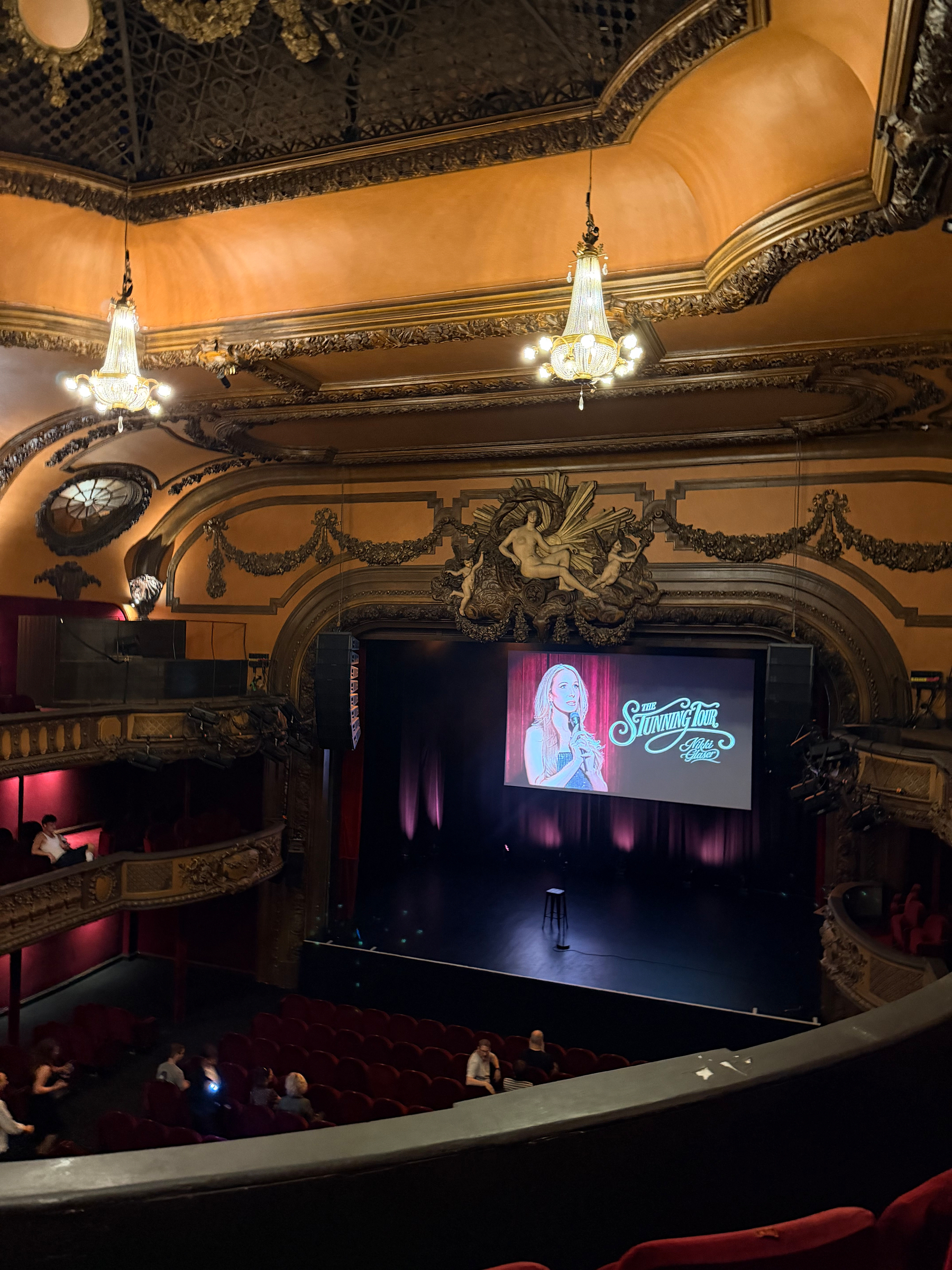An ornate theater with a grand stage setup features a microphone stand and a backdrop promoting Stunning Tour with Nikki Glaser, as a small audience gathers.