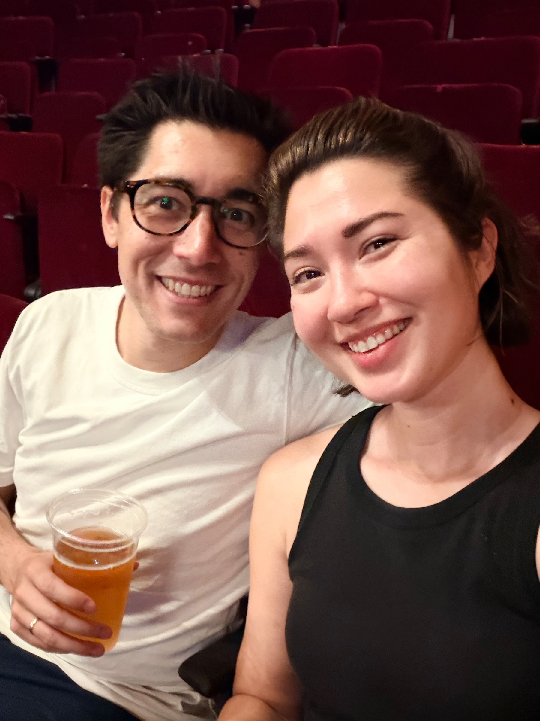 A smiling couple is seated in a theater or auditorium, with one holding a cup of beer.
