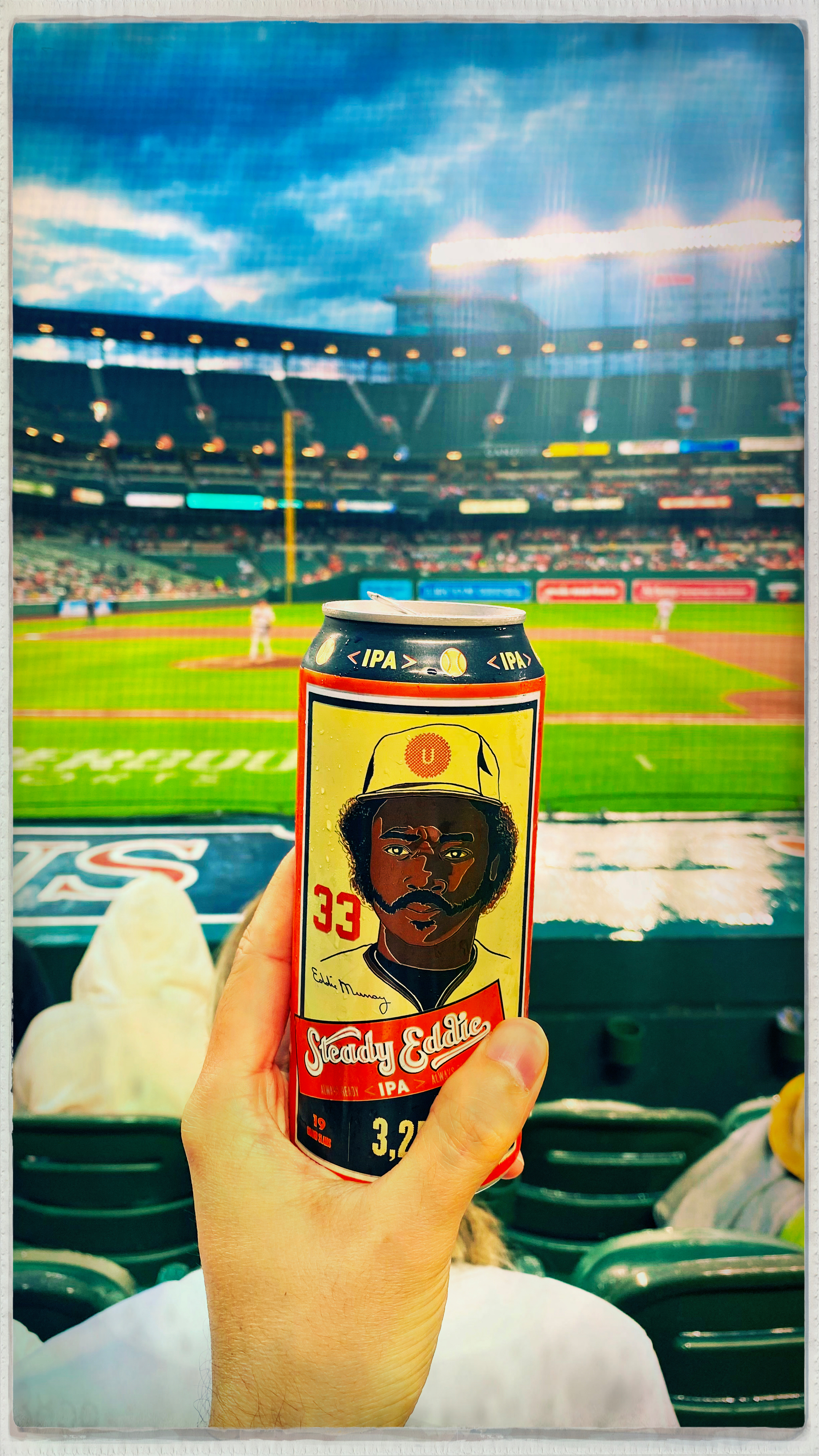 A person is holding a can of IPA beer with a baseball-themed design in a sports stadium.