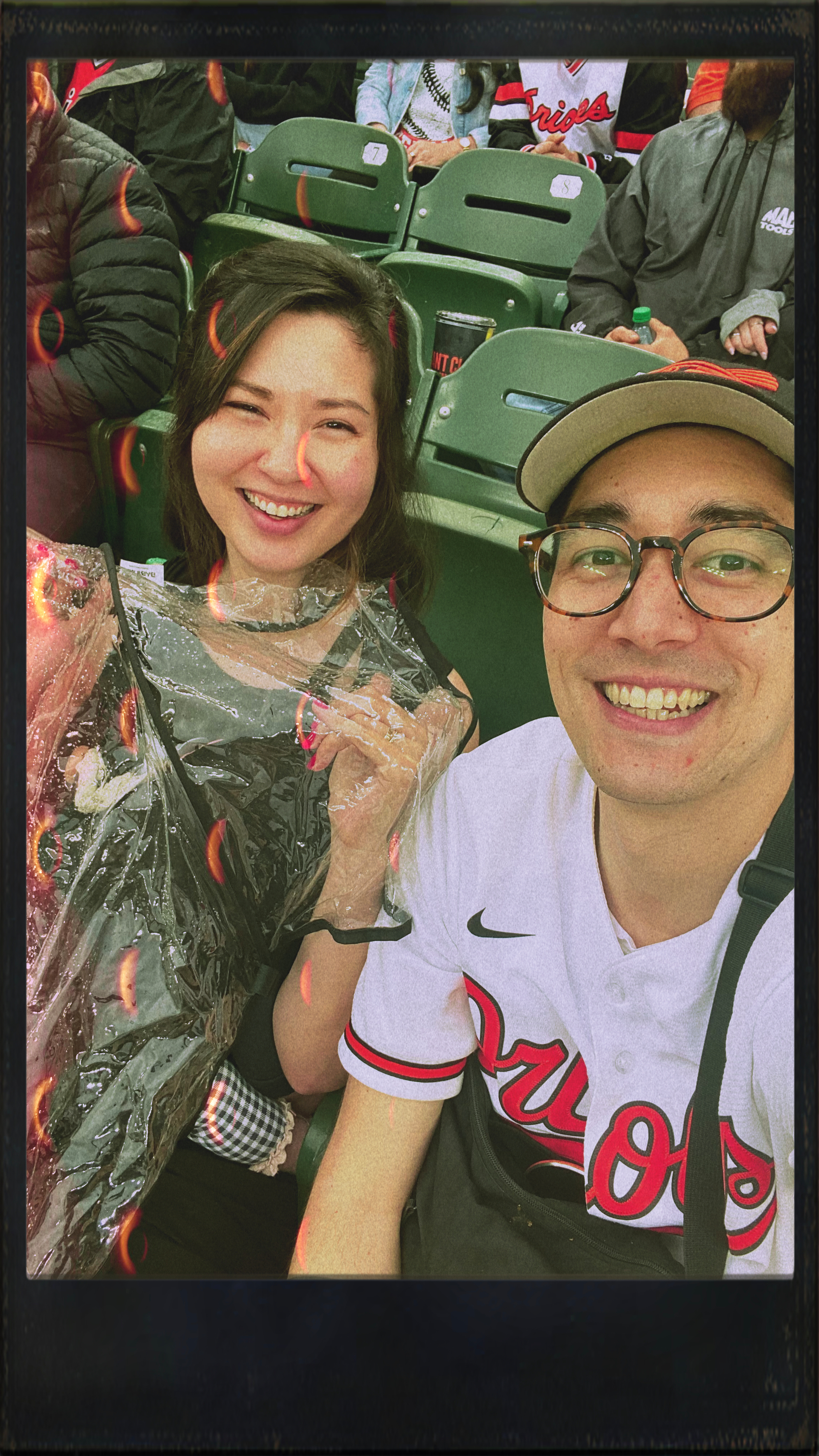 Two people are sitting in stadium seats, smiling, with one wearing a raincoat and the other wearing a baseball cap and jersey.