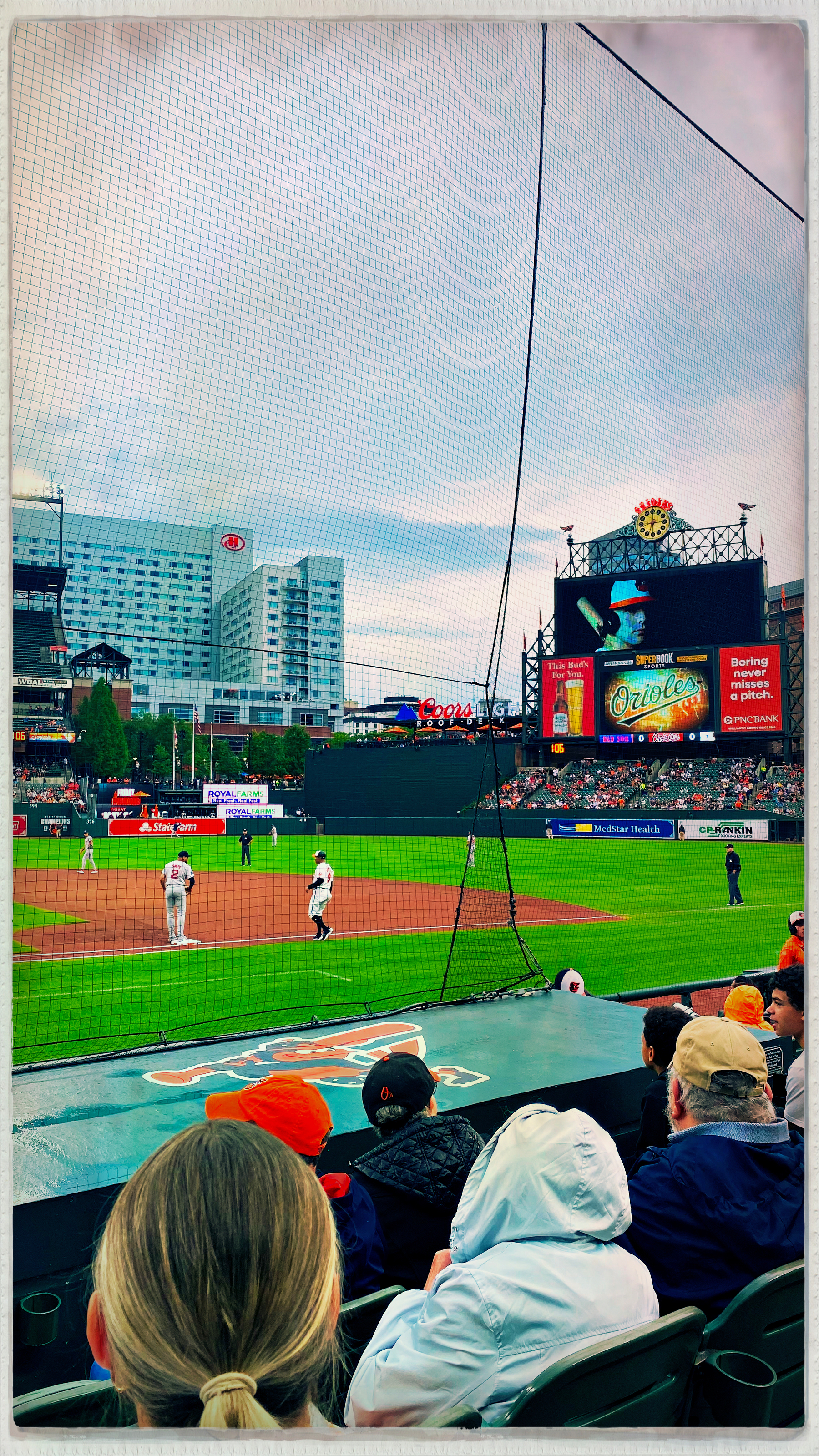 Fans watch a live baseball game from the stands, with the field and scoreboard visible in the background.