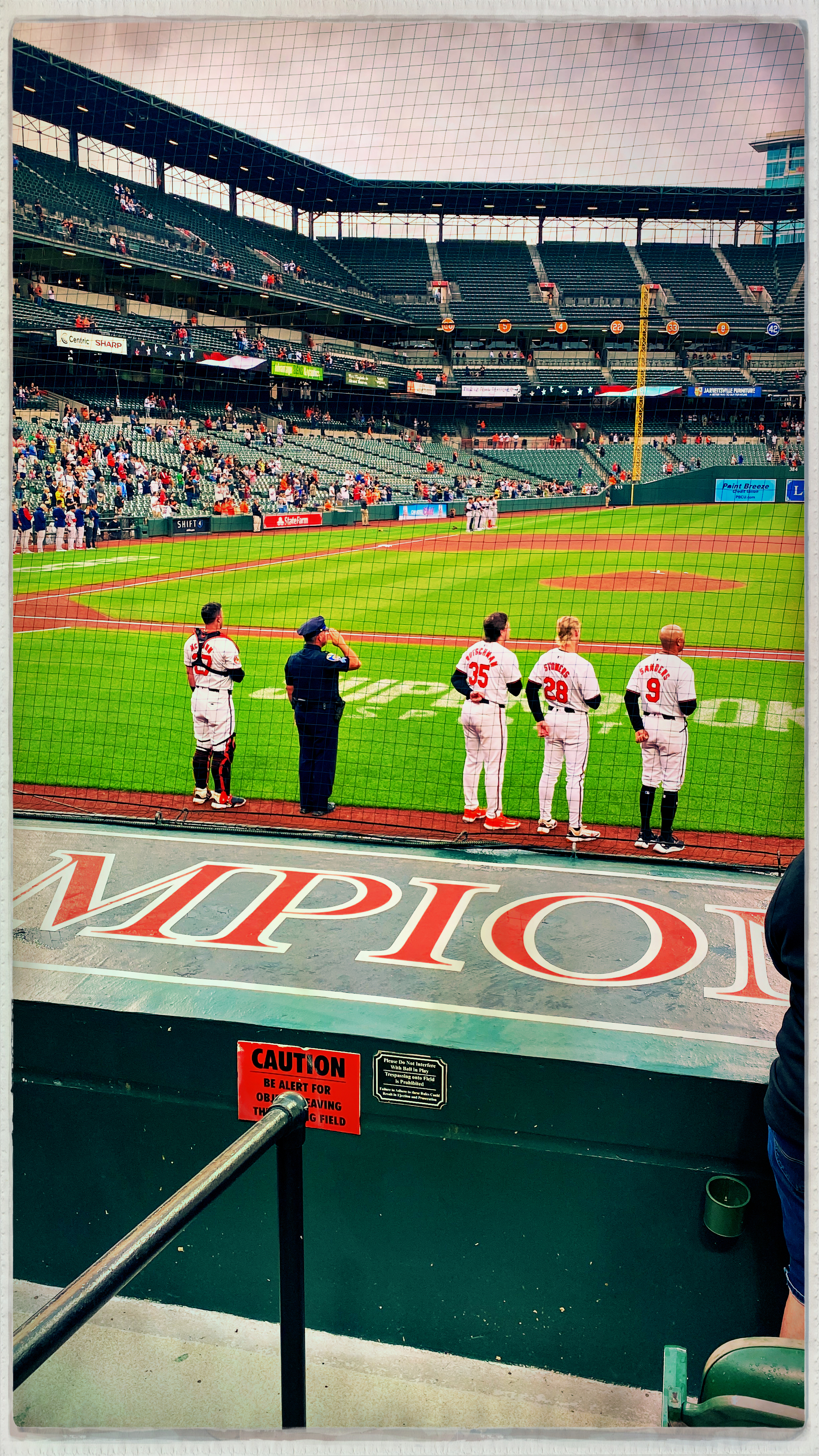 Baseball players are standing near the field while spectators watch from the stands.