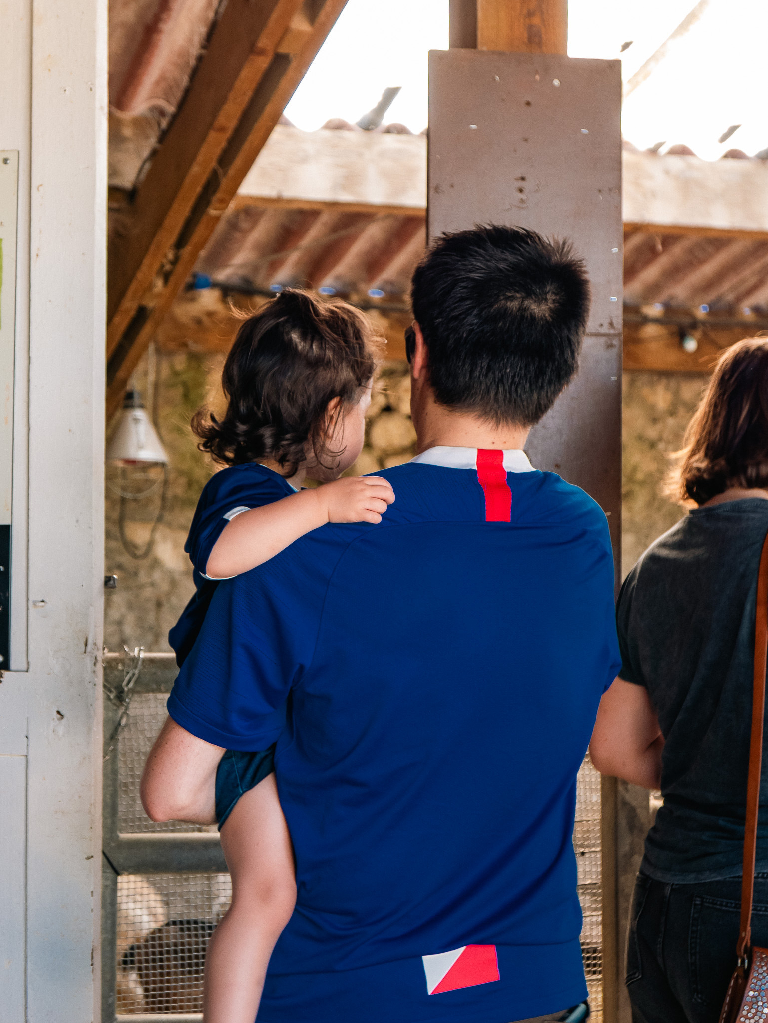 Auto-generated description: A man wearing a blue shirt is carrying a child on his left arm while standing indoors near another person.