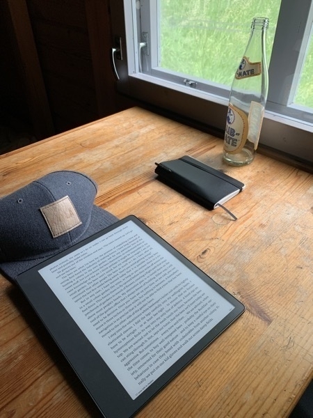 A photo of table inside a cabin, on which are laying a few items: a base cap, an empty soda bottle (club mate), an ebook reader and a notebook with a strap holding it closed and a pen tucked inside it