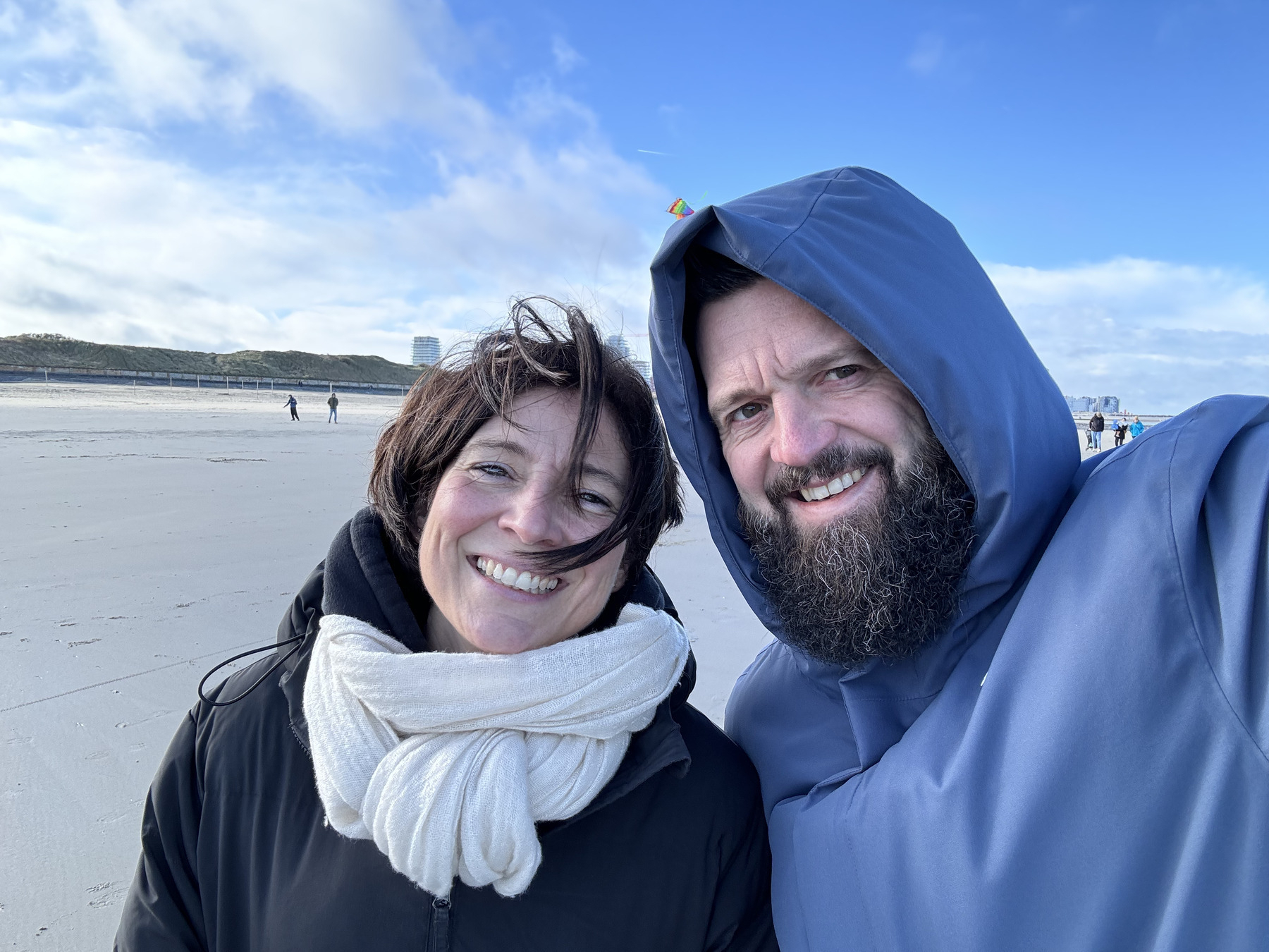 Two smiling adults in hooded jackets take a selfie on a sandy beach with scattered people and a dune landscape in the background.