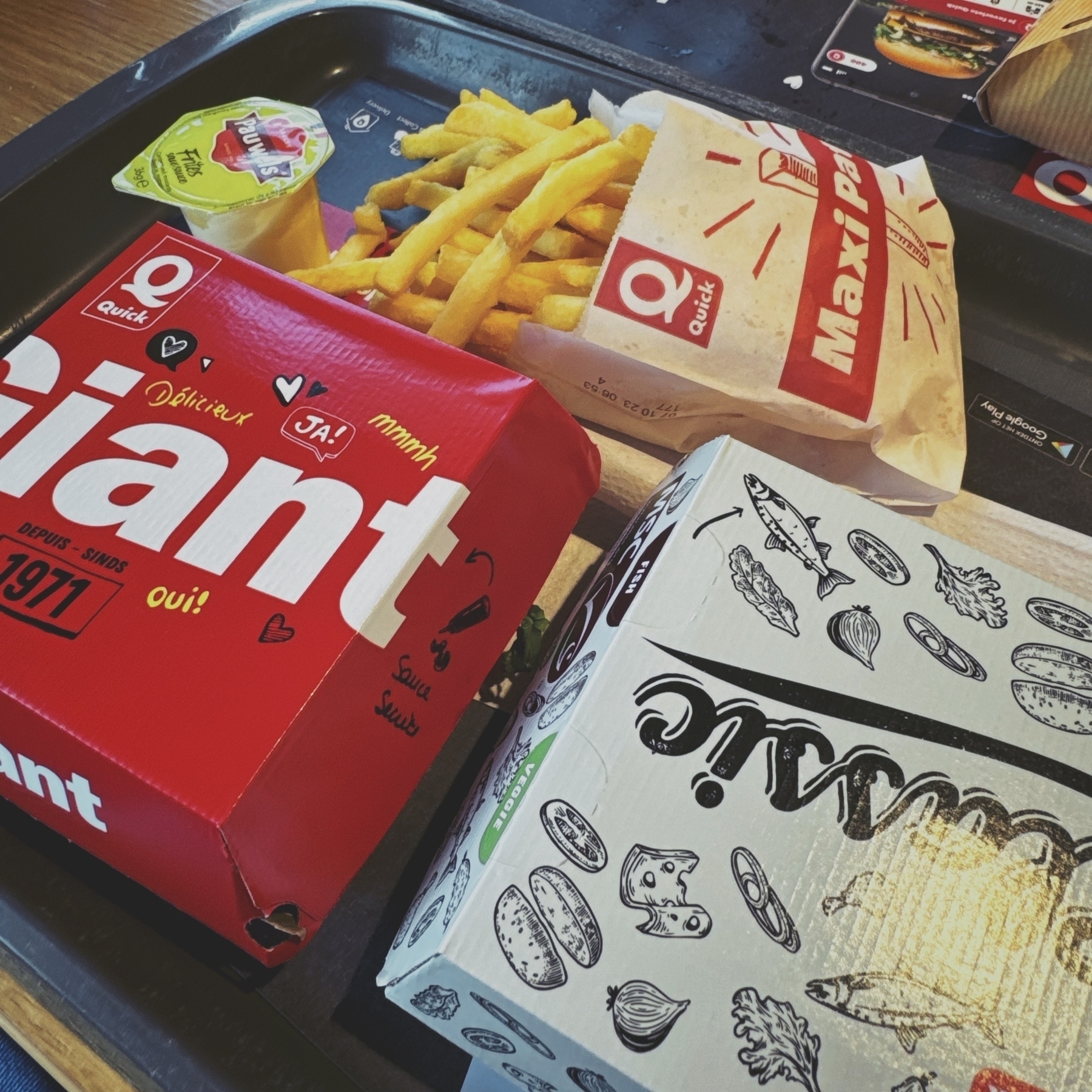 Fast food items on a tray featuring a burger box, fries, a sandwich wrapper, and a small cup of sauce, in a casual dining setting.