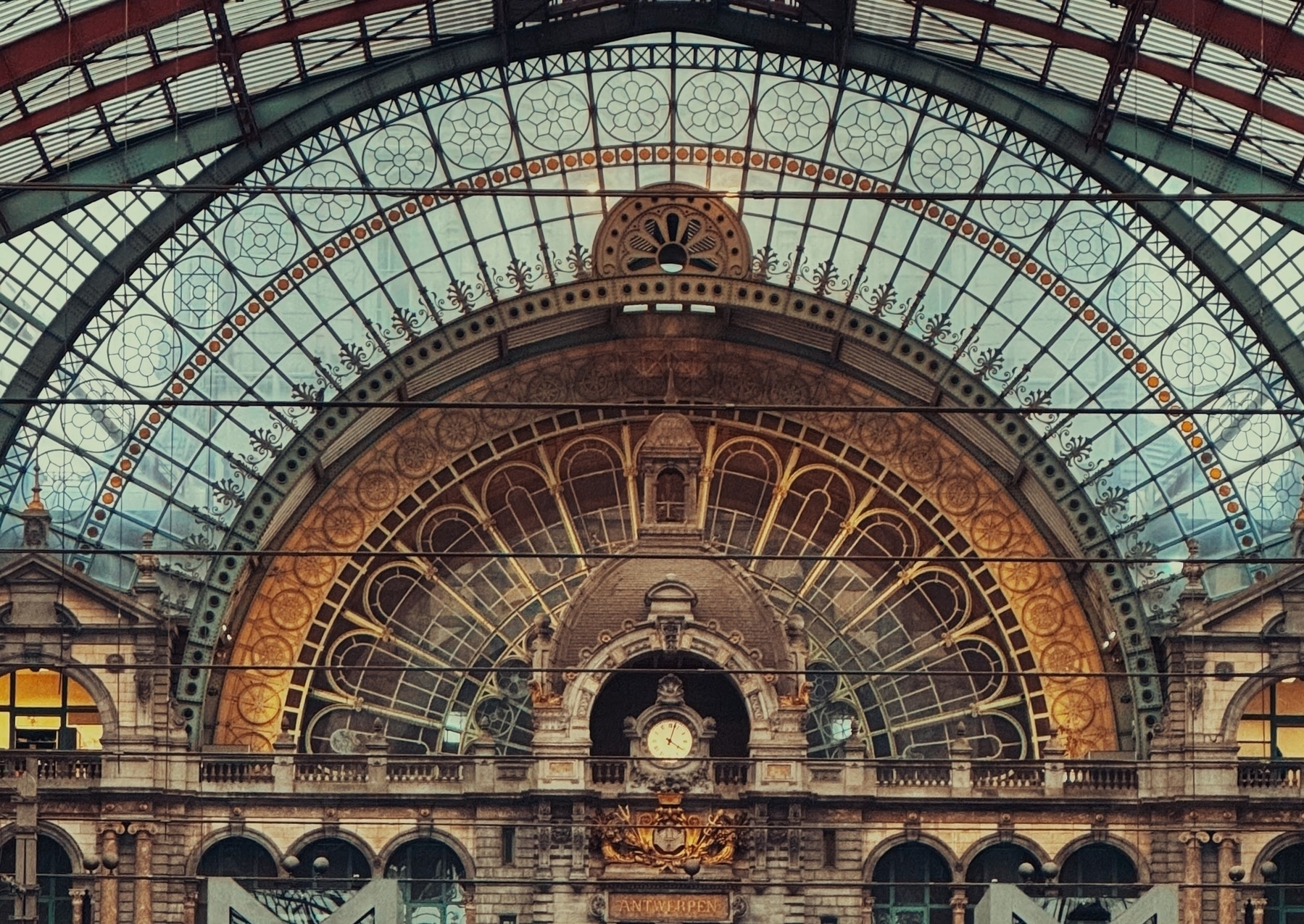 An ornate train station interior with arched glass ceilings and detailed stonework, featuring a central clock and the word “ANTWERPEN” on a decorative backdrop.