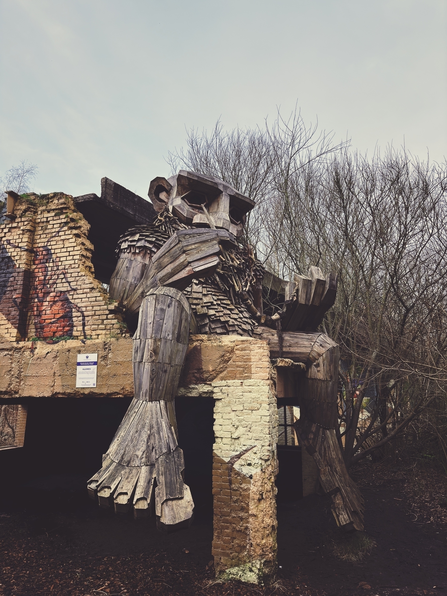 A wooden sculpture resembling a humanoid giant emerging from ruins, against a backdrop of leafless trees and a clear sky.