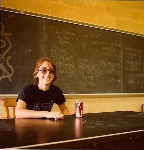 A young Brent sitting at a desk with a Coke can. Brent has long hair; he’s wearing slightly tinted glasses and a Space Invaders T-shirt. In the background is a chalkboard with some equations.
