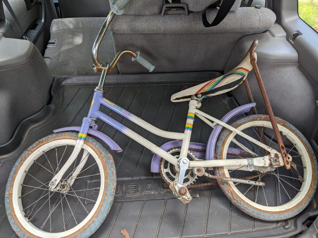 Rusty and neglected children's bicycle with white and lavender paint, rainbow accents and banana seat.