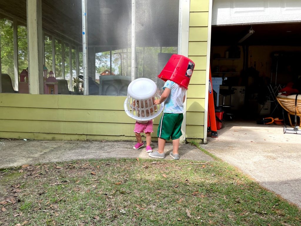Two young children.  One with a red bucket on his head and one with a laundry basket on her head.