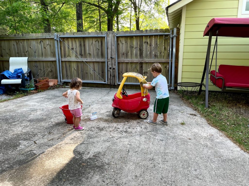 Young boy washing a plastic toy car with a sponge.  Young girl next to a red bucket.