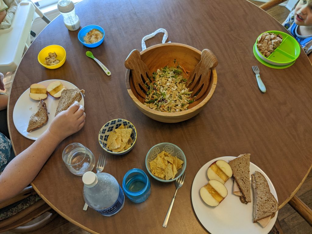 Image of dinner table with salad bowl, plates with sandwiches and apples, and smaller bowls for children.