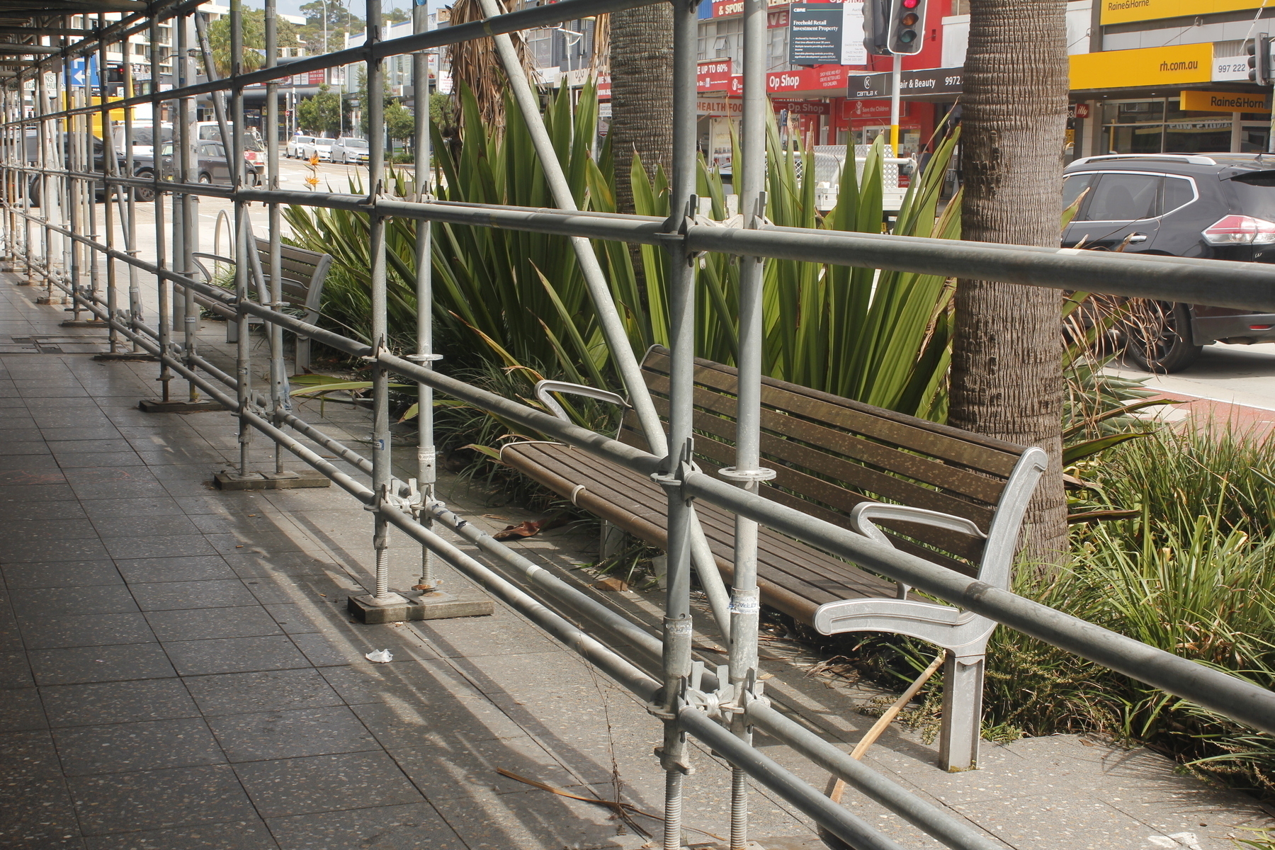 Beside a busy road, building scaffolding begins in the right foreground and stretches into the distance, top left corner. The scaffolding stands at the edge of a tiled footpath, and immediately outside it is a bench, with metal frames and arm-rests and brown timber slats. There’s very little room to sit comfortably. Against the back of the bench is a planted area with tall grasses, trunks of palm trees and tall, dense, spiky green semi-succulents.