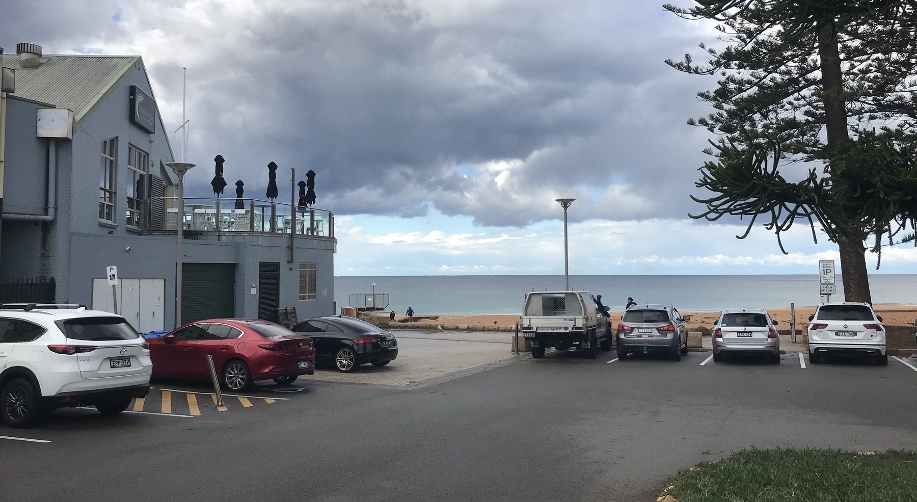 Looking across a carpark with a grey, gabled building on the left and a Norfolk Pine tree on the right, towards a beach, a dark grey sea with a flat horizon, and a sky with two contrasting aspects. For a short distance above the horizon the sky is pale blue with fluffy white clouds. Above this hang dark stormclouds, reaching all the way to the top of the photo. The grey building on the left has a balcony with five black umbrellas standing folded against the dark grey clouds. Two boys in black are hurrying up the beach towards the carpark.