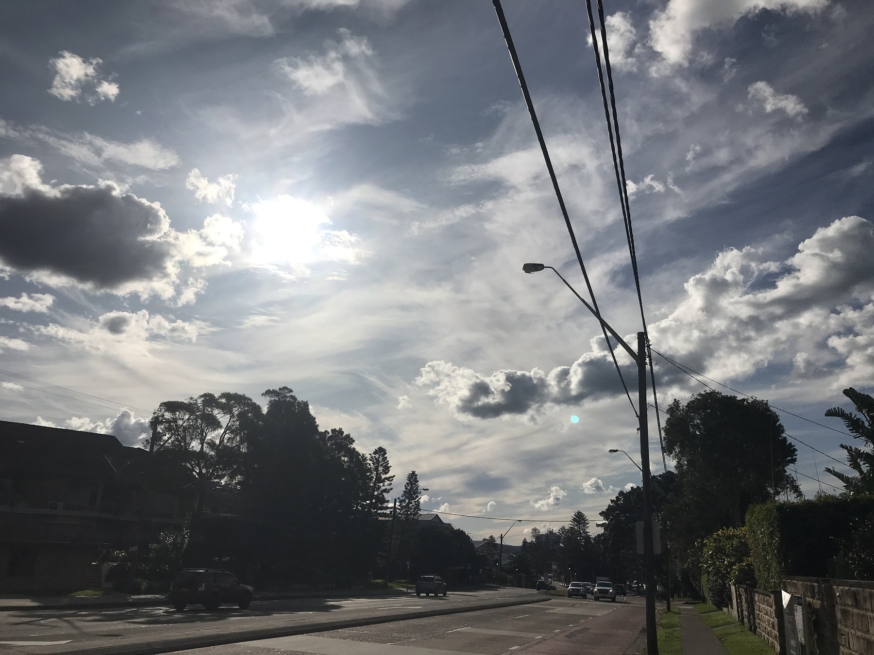 The sun is a hot white glow behind a bank of cloud, in a late afternoon sky alive with cloud formations. Deeply shadowed cumulus hangs above the ground like storm clouds, and higher up there are wisps, slashes and trails of white and grey against grey blue rivers of sky. The foreground is a main road edged with suburban blocks, receding into the distance. One side is in deep shade. On the other stands a telegraph pole, in crisp silhouette, with a street light arching over the roadway and three overhead cables exiting the top of the frame. Tall trees rise in silhouette against the glare, and a small blue disc of lens flare hangs like a second sun.