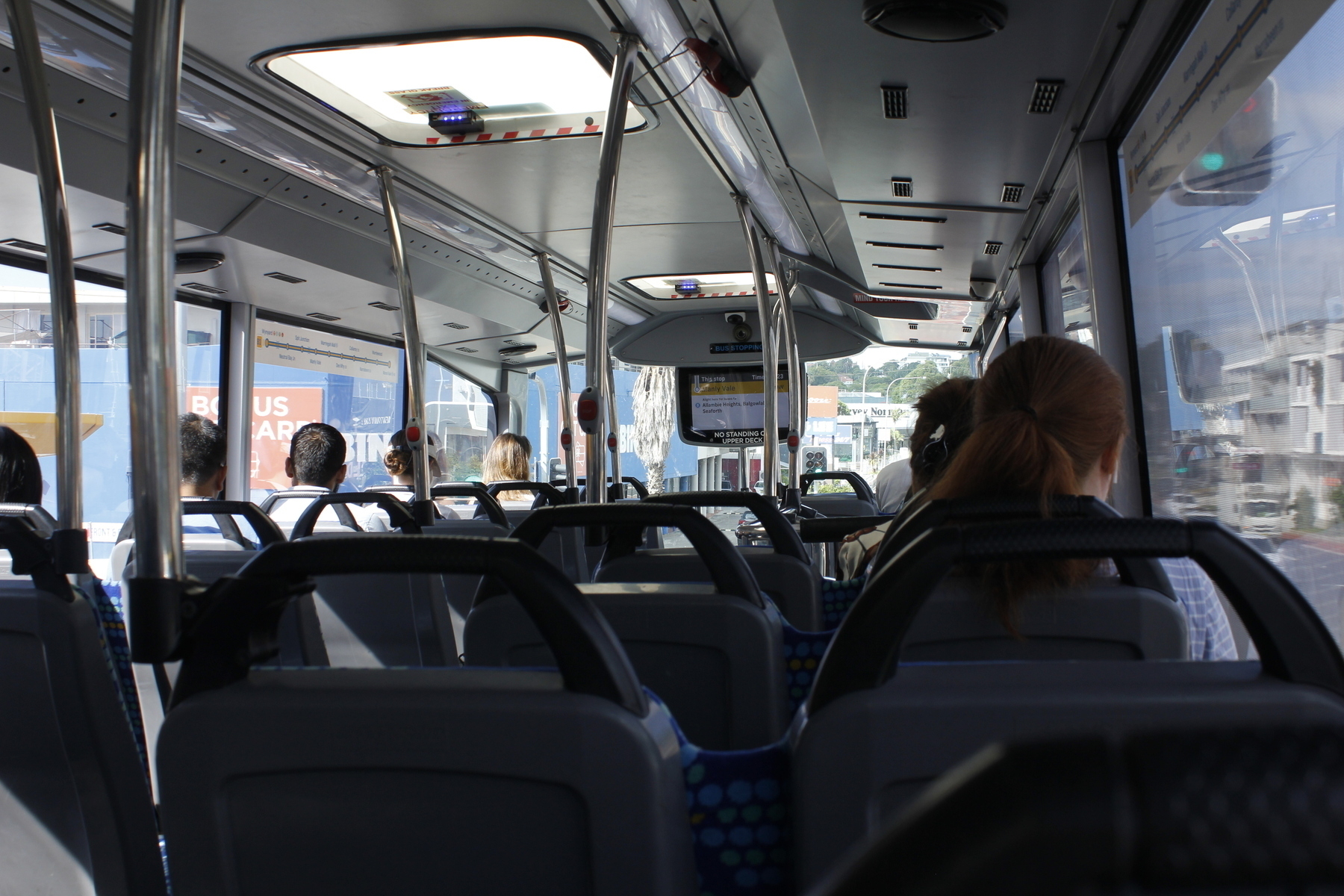 A view from near the back of the top deck of a double-decker bus, looking forward at the backs of five passengers’ heads on the left and two on the right. A screen at the front says ‘This stop: Manly Vale’. Outside are shop signs, apartments and traffic lights.