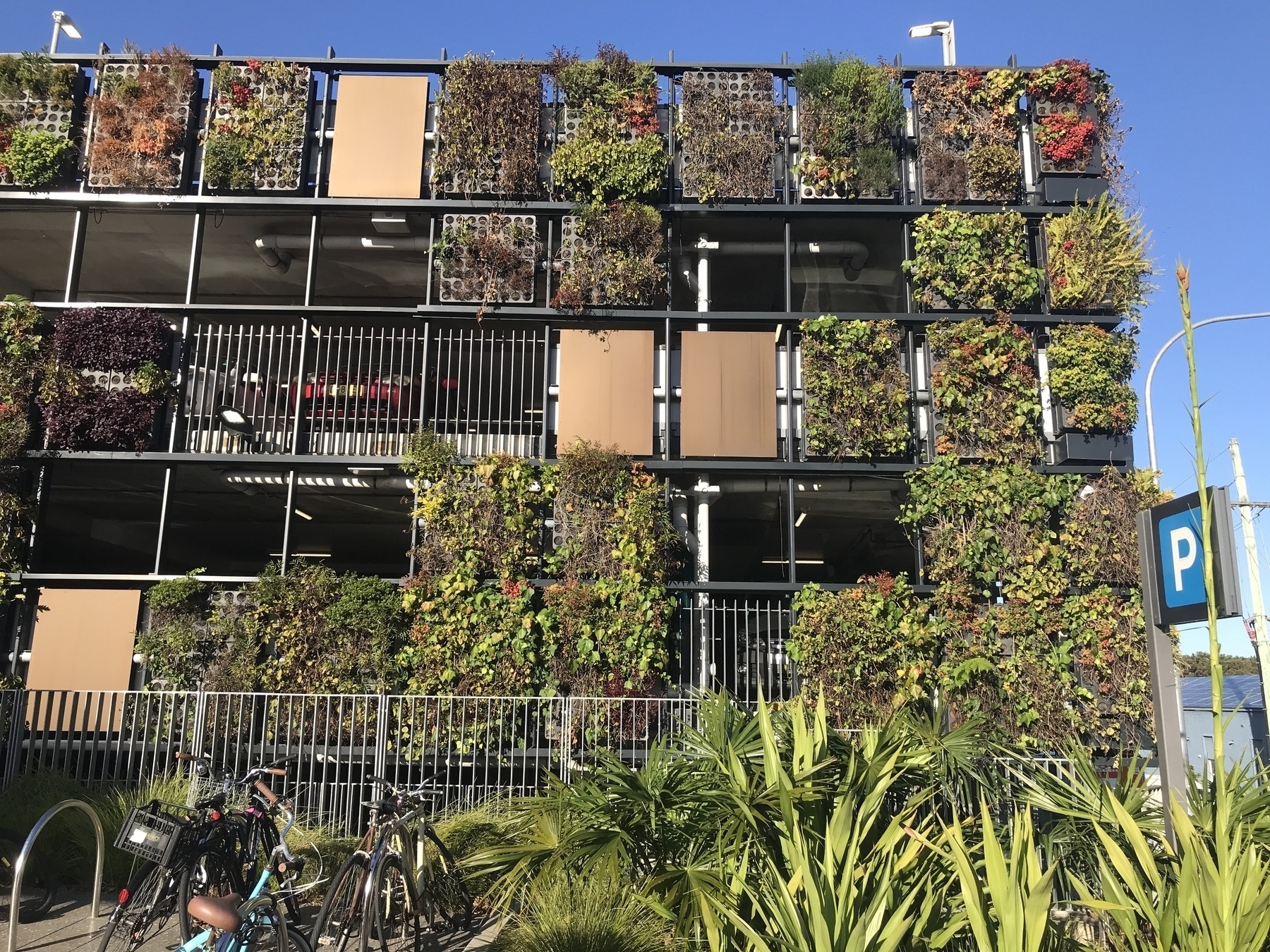 Part of the front of a three-storey carpark seen from the footpath. The carpark’s facade is composed of squares and rectangles, two levels per storey. Some are open to reveal the interior. Some are enclosed by vertical rails, some by solid panels, and many others by thriving plants grown in the holes of concret panels or in tiered garden  boxes. On the path is front of the carpark is more greenery and a bicycle parking space.