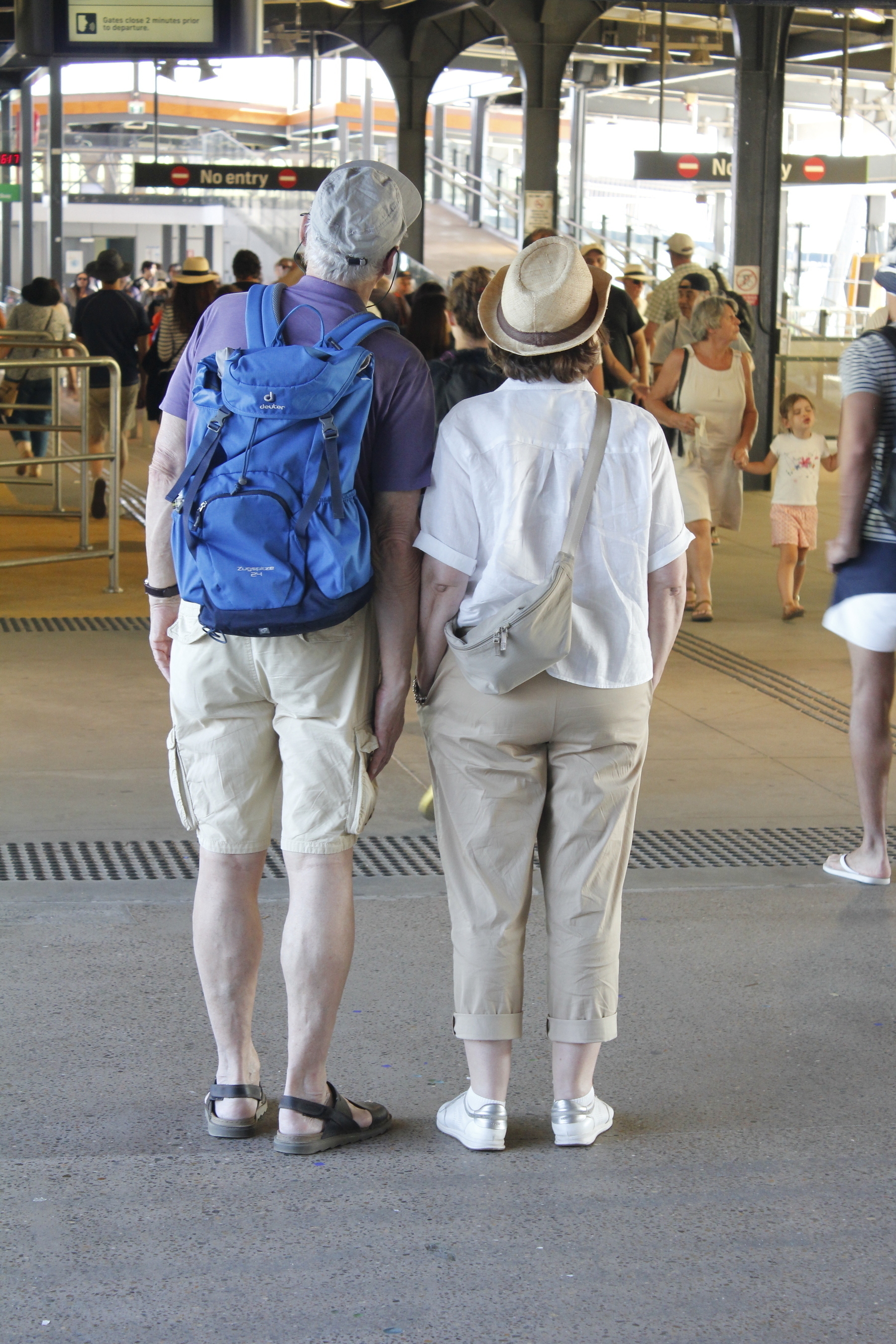 A middle-aged couple seen from behind, standing side by side, heads raised and angled in the same direction to read the ferry departure times. A crowd of disembarking passengers walks towards them.