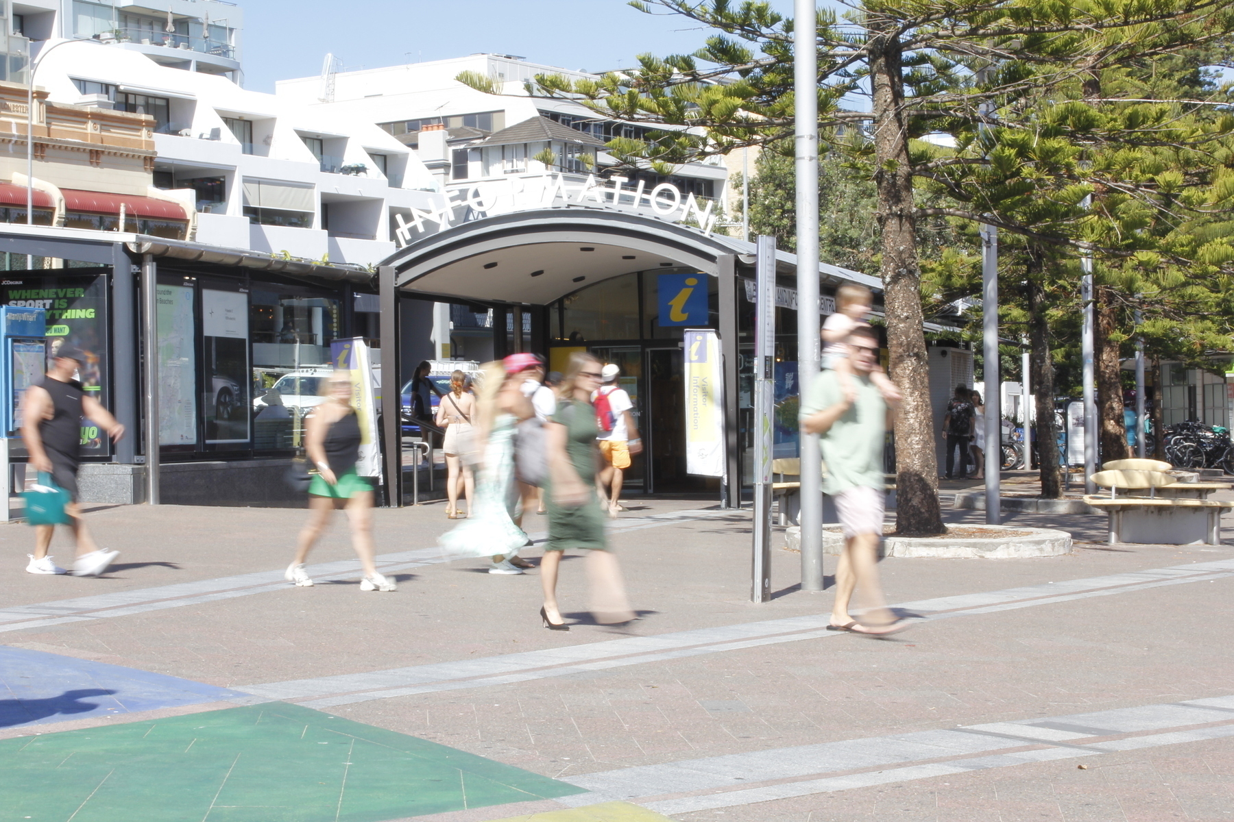People walk from left to right across a sunlit plaza, looking blurred because the long exposure has captured their movement. One foot of each walking person is clear, not blurred, because that foot was not moving when the shutter opened. Everything else around the walkers is clear and in focus, like figures standing in the background, and an arched entrance saying Information.