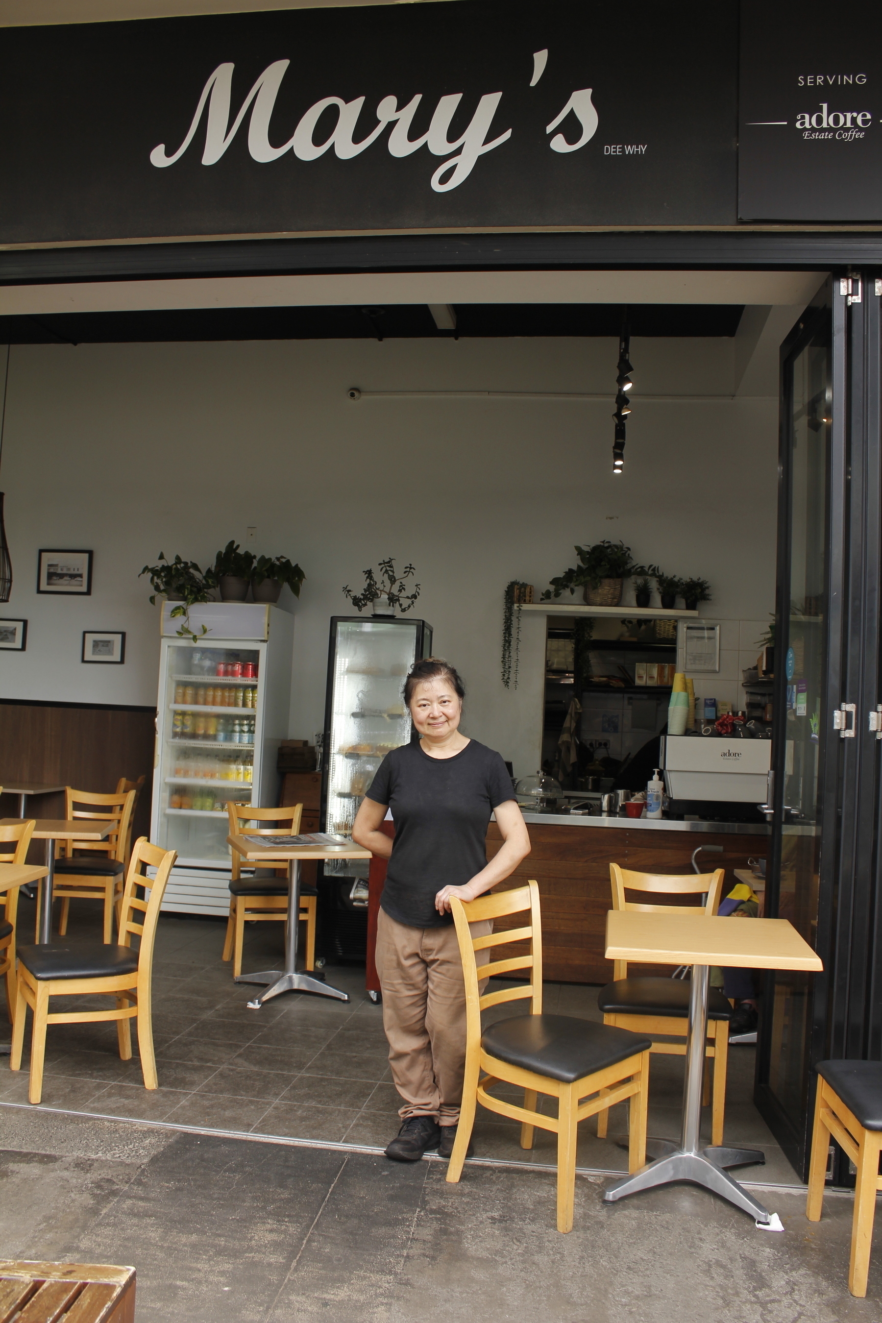 A woman stands just inside an open-fronted cafe, resting one hand on the back of a chair and smiling at the camera. She wears a black t-shirt, brown trousers and dark brown boots, and has one knee bent. Inside the cafe are tables and chairs, two tall fridges against the far wall, the serving counter and, behind it, a doorway leading into the kitchen. The floor is tiled, and glass doors are folded back against the right-hand side. A sign above the entrance says “Mary’s Dee Why” and “Serving Adore Estate Coffee”.
