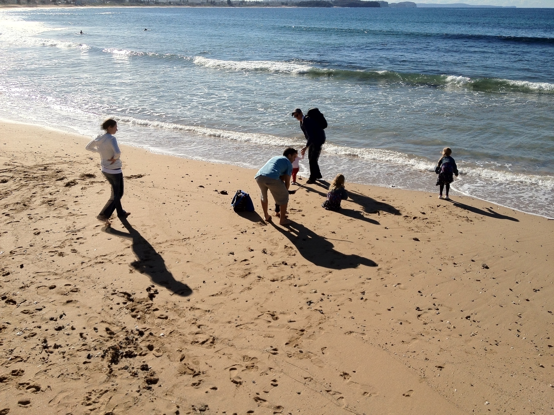 A group of adults and children cast long shadows as they fossick on a beach. Everyone is dressed for Winter, in jumpers and long trousers, except for a man in shorts and t-shirt who bends down to support a child, a blue backpack on the sand beside him. A second man, almost in silhouette at the water’s edge, fully dressed and carrying a backpack, leans forward holding the hand of a toddler in a pink top, red pants and trainers. The man’s heel is lifted, with water flowing beneath. Nearby are two small girls with long blonde hair. They’re dressed alike, and may be twins. One crouches down on the sand, hair loose, and the other stands facing the sea with her toes almost touching the waves. She wears a jacket with long sleeves, a knee-length dress, dark tights and white trainers. Her hair is in a plait. One arm lifts slightly, as if she wants to reach out. A woman walks down the sand towards the group, hair up, hands on hips. She wears long pants and a white hoody, but her feet are bare.
