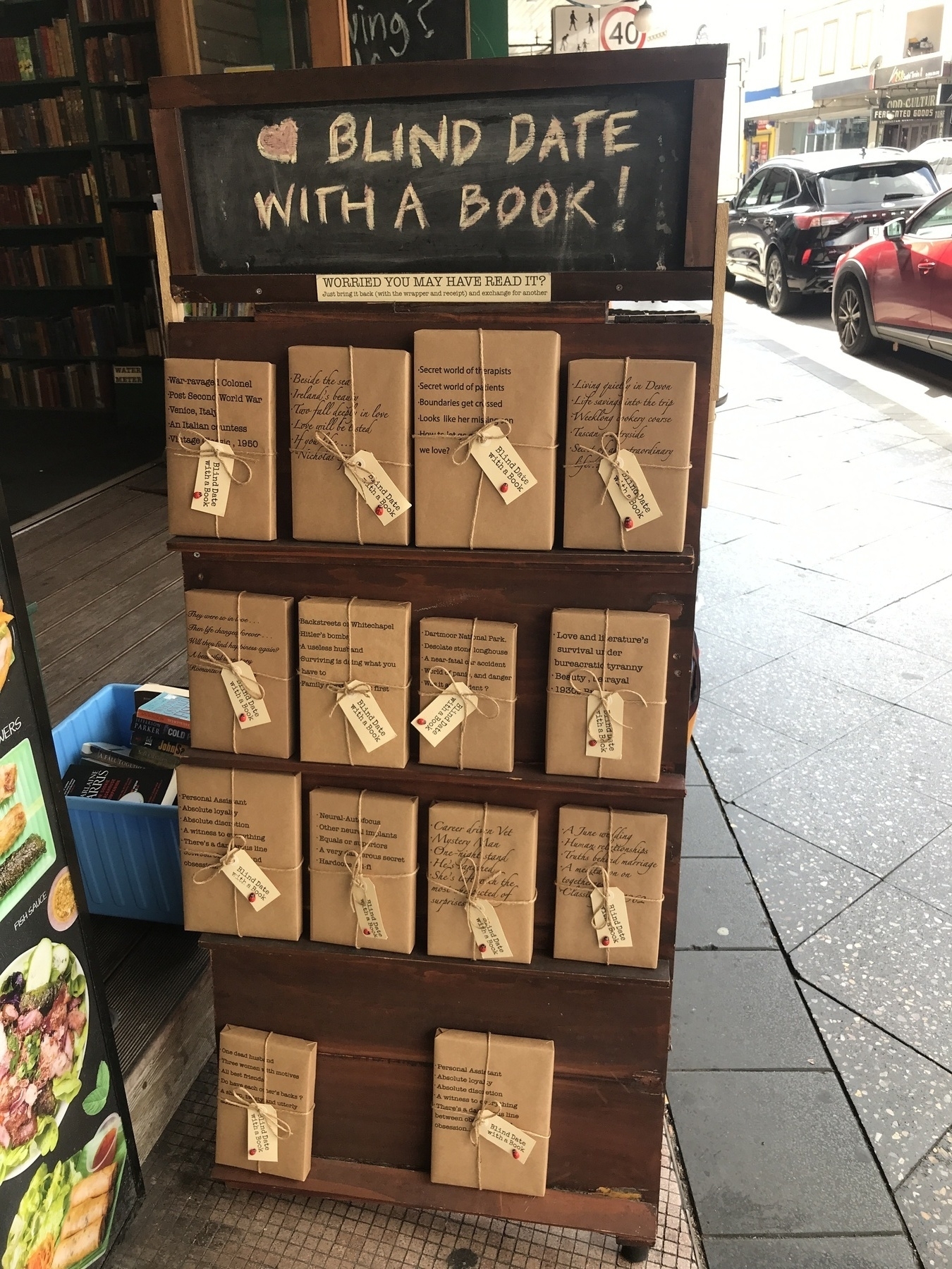 A timber stand on a footpath by a doorway, with parked cars in the background. The stand has four narrow shelves, and each shelf holds books facing outwards, wrapped in brown paper and tied with string. The top three shelves have four books each, and the bottom shelf has two. Key phrases about each book are written on the front, like ‘Career driven vet’, ‘A June Wedding’, ‘Backstreets of Whitechapel’ and ‘War-ravaged Colonel’. A chalkboard above the stand says: ‘Blind Date with a Book!’ with a heart beside it. A “sticker beneath that says: ‘Worried you may have read it? Just bring it back (with the wrapper and receipt) and exchange for another.