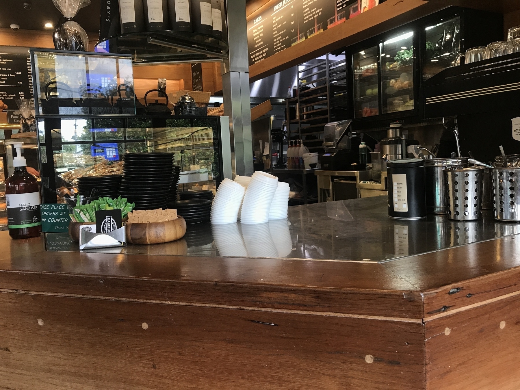 On a highly reflective cafe counter crowded with lids, business cards, wooden bowls, condiments, hand sanitiser and stainless steel containers, four close-packed piles of white throwaway coffee cup lids curve gracefully sideways, like plants reaching for the light. They are mirrored in the counter top.