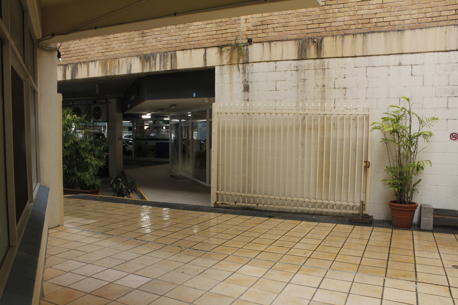 A wide rectangular space in a wall opens through to a shadowy realm of plants, pillars, cars, fluorescent lights and a walkway passing glass doors. Before the wall is a shopfront on the left and a floor of square tiles. The tiles closest to the wall are wet and shiny. A wide steel gate folds back against the wall, next to a tall potted plant.