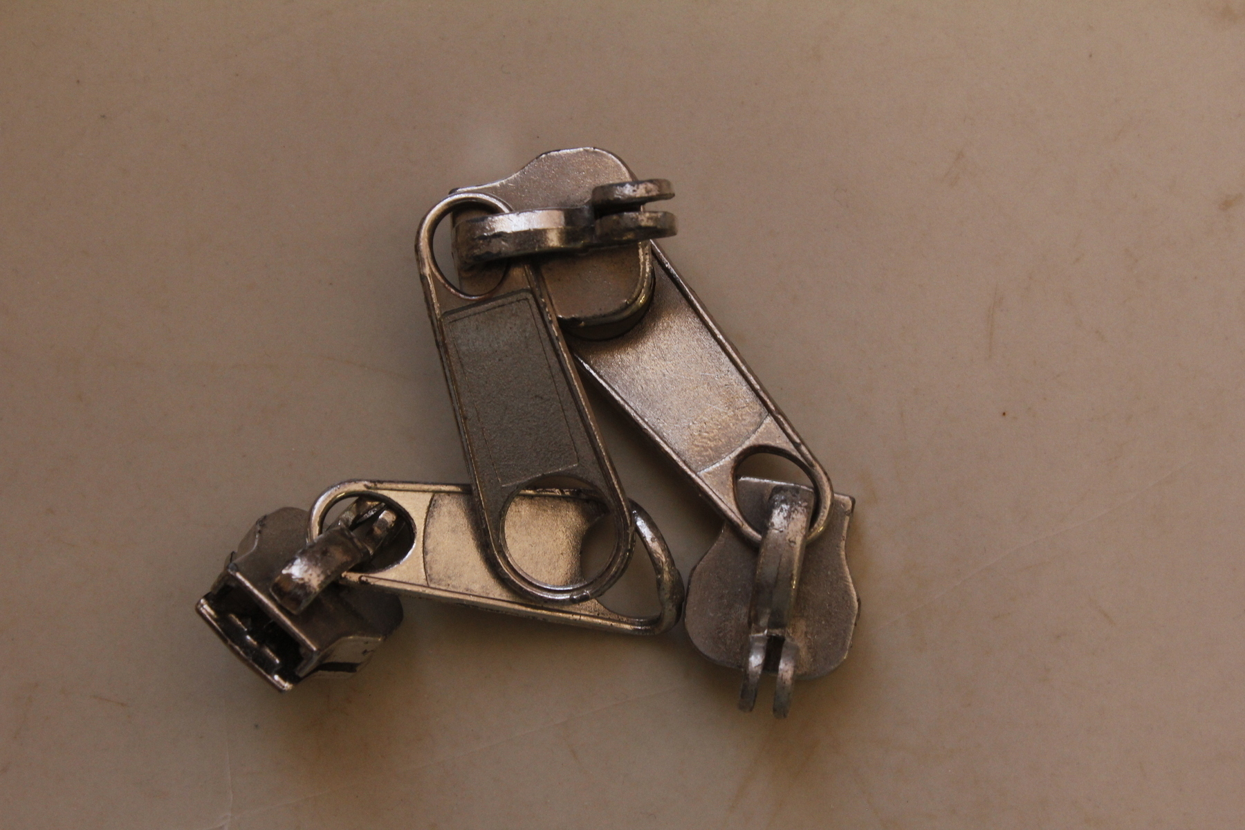 Three shiny zipper handles, no longer attached to zippers, lie jumbled together on a soft, mottled brown background. The camera has zoomed in so the zipper handles seem big, and detailed. Illumination is from the side, lighting some surfaces and throwing others into shadow.