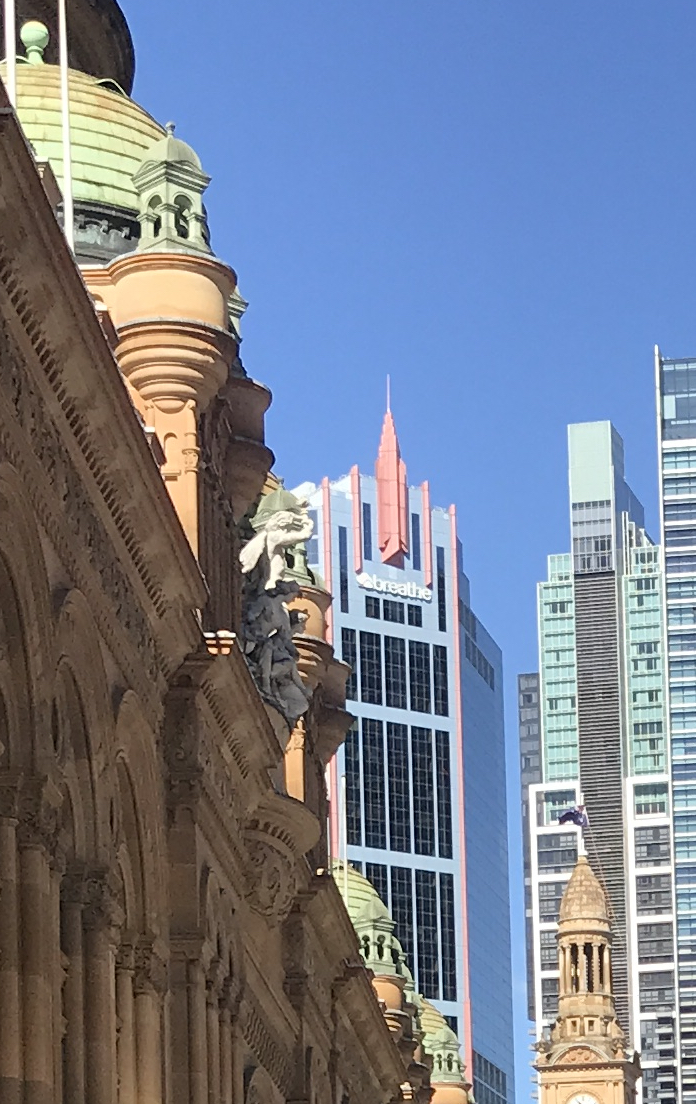 Old and new buildings are juxtaposed against each other, and against a blue sky in the sunshine. On the left is the Queen Victoria Building with its sandstone walls and sculptures and its green copper domes, overlaid on the blue exterior of a tall new building with a pink spire, pink trim, and blue-edged blocks of dark windows. To the right of this, almost as tall and almost touching it, is a grey and aqua green facade surmounted by a narrow block of windows and flat green wall, echoing the pink spire; and down low, against this building, stands the sandstone clock tower of the Sydney Town Hall, dwarfed by its modern neighbours.