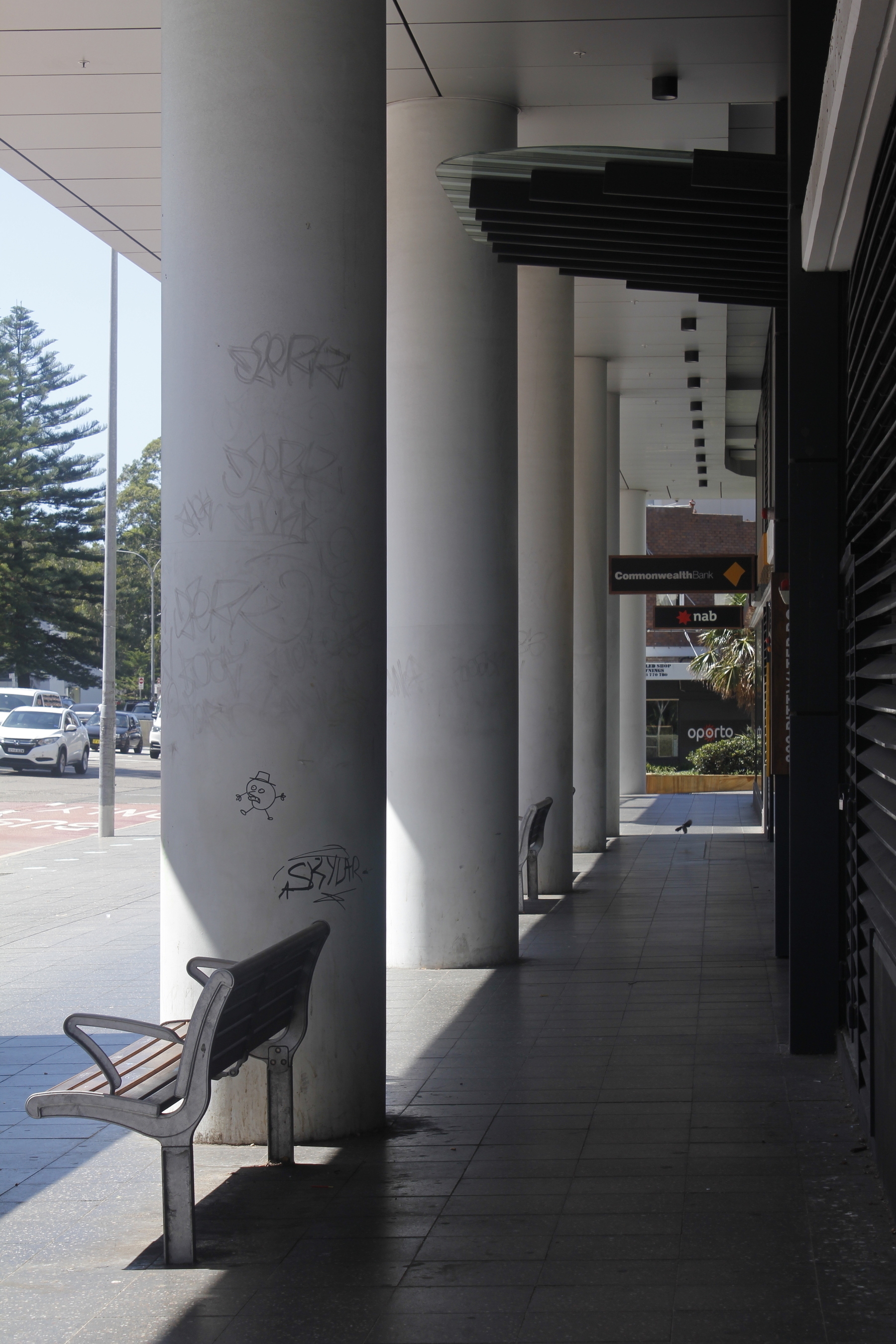 A line of very thick white columns runs into the middle distance, with a roof projecting out over the tops of the columns, on the left. To the right of the columns is a long stretch of tiled footpath and the external wall of a commercial building, both in deep shade. To the left of the columns is hot, bright, glaring sunshine and a glimpse of cars on a main road with trees in the background.