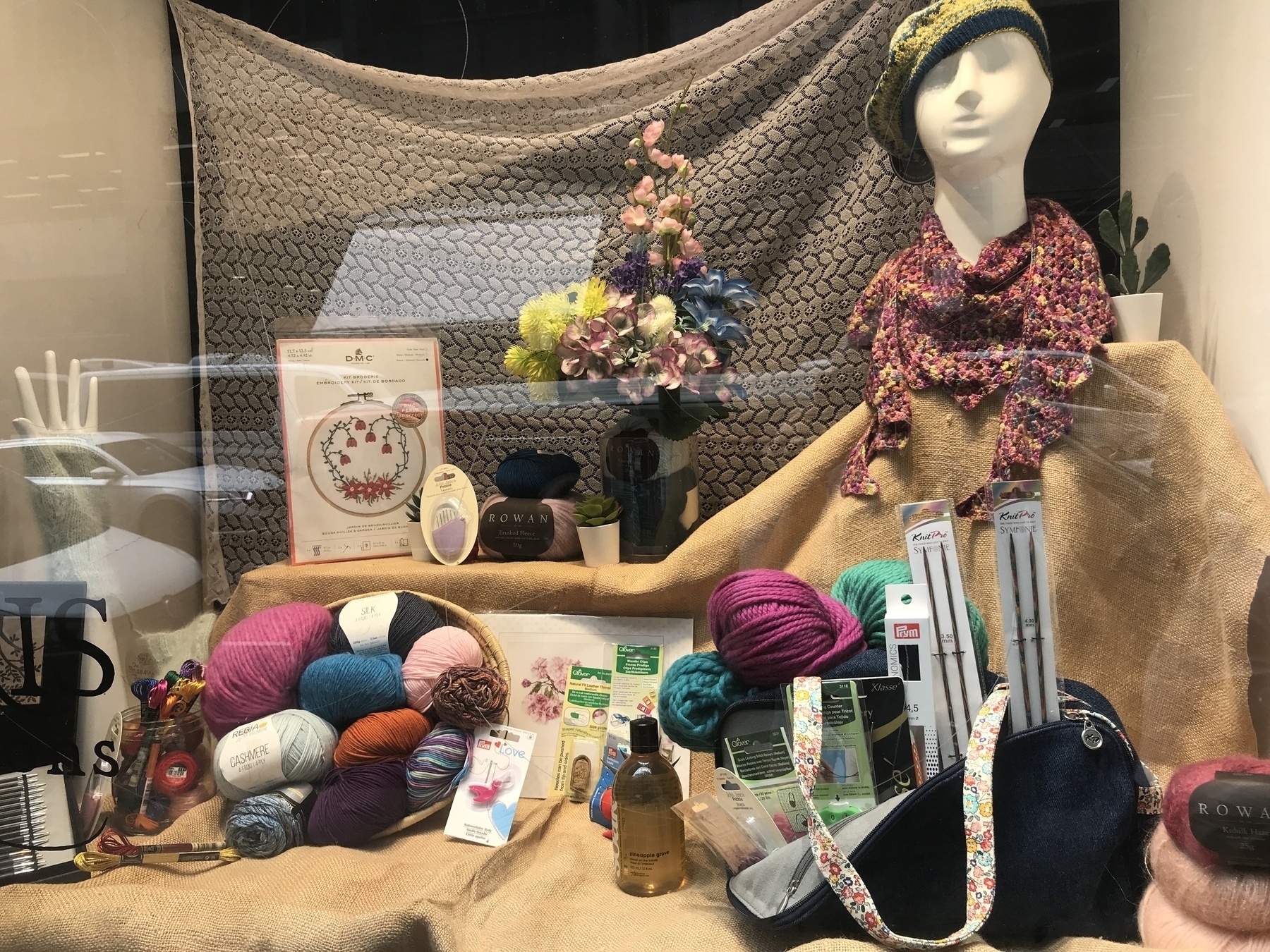 The window display of a needlecraft shop. Against a background of crocheted blanket and plain hessian sacking, colourful balls of wool tumble from basket and bag amid examples of embroidery kits, knitting needles, bags, embroidered straps and labels, and bundles loops of thread. A tall arrangement of flowers fills the middle, with a tiny small potted succulent at its base, and on the right is a mannequin’s head and shoulders wearing loops of thick, knitted scarf in autumn colours, and a floppy knitted beret in black and yellow. At the back, on the left, an upstretched mannequin’s hand emerges from a green knitted wrist-warmer.